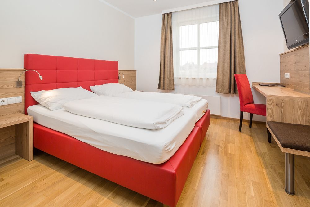 Moderná hotelová izba s červenou posteľou a písacím stolom.