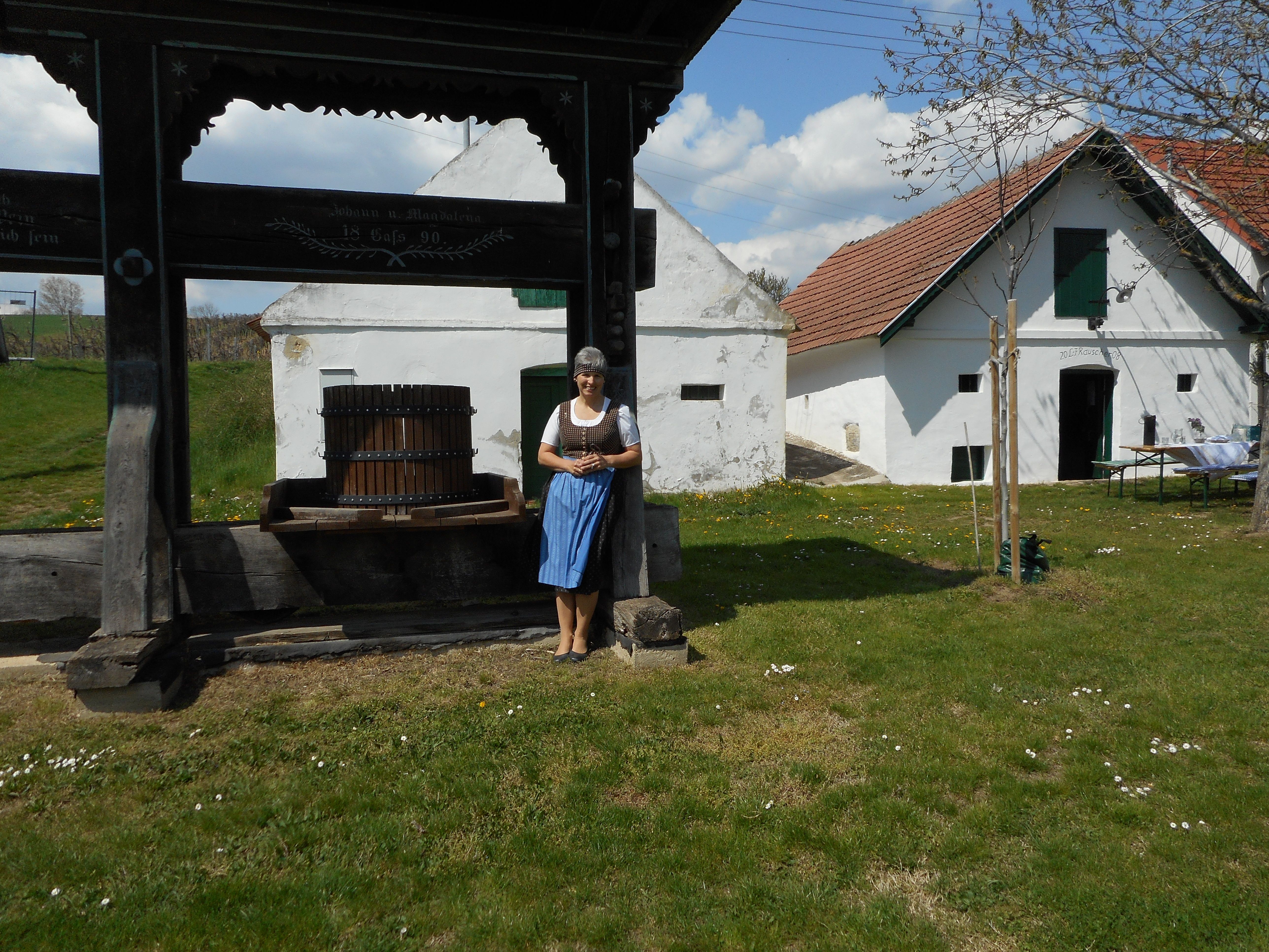 Žena v tradičnom odeve stojí vedľa starého lisu na víno pred historickou budovou s bielymi stenami a červenými strechami.