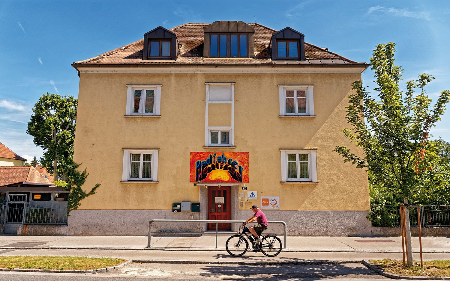 Žltá budova s červeným vchodom a nápisom graffiti. Cyklista prechádza okolo. Modrá obloha a stromy v pozadí.