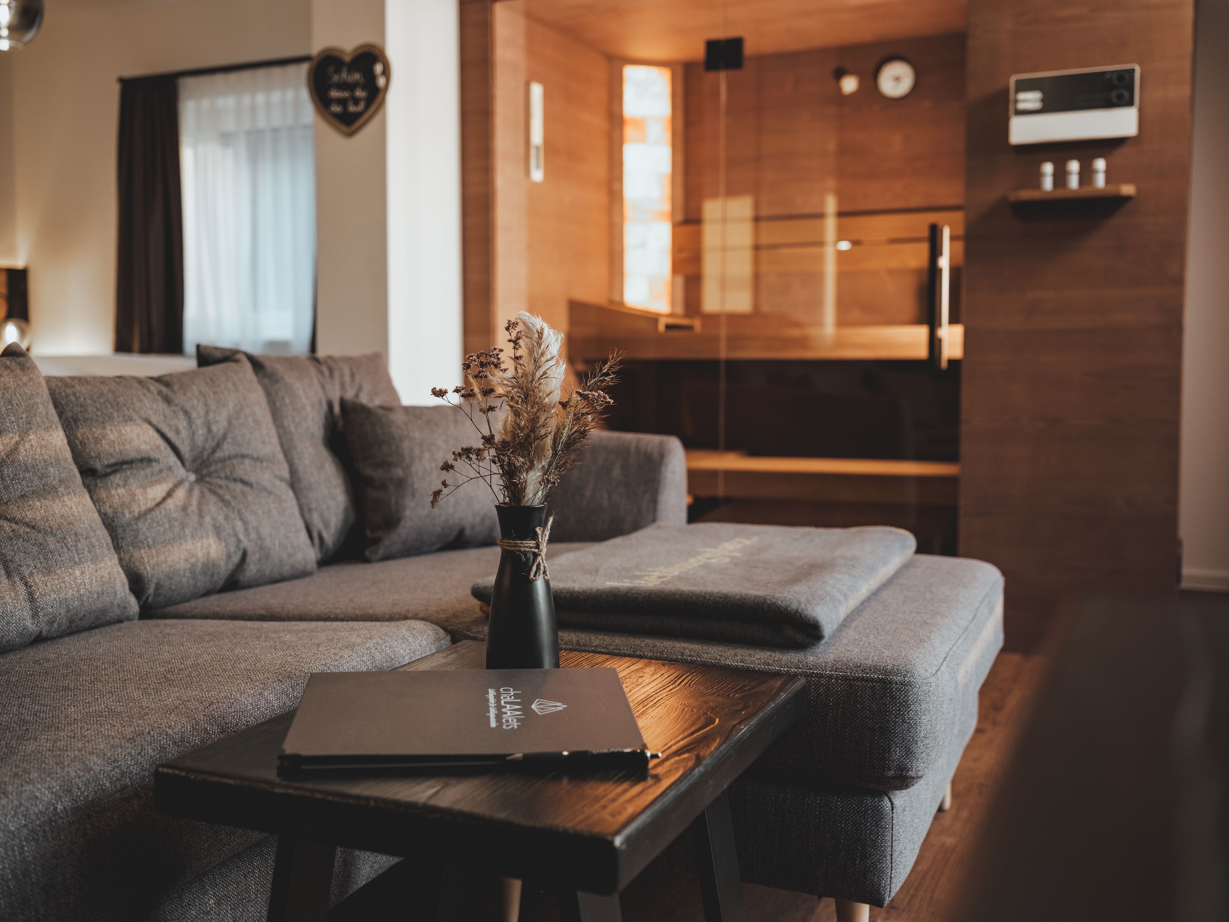Útulná obývacia izba so sivou pohovkou, dreveným stolom a saunou v pozadí.