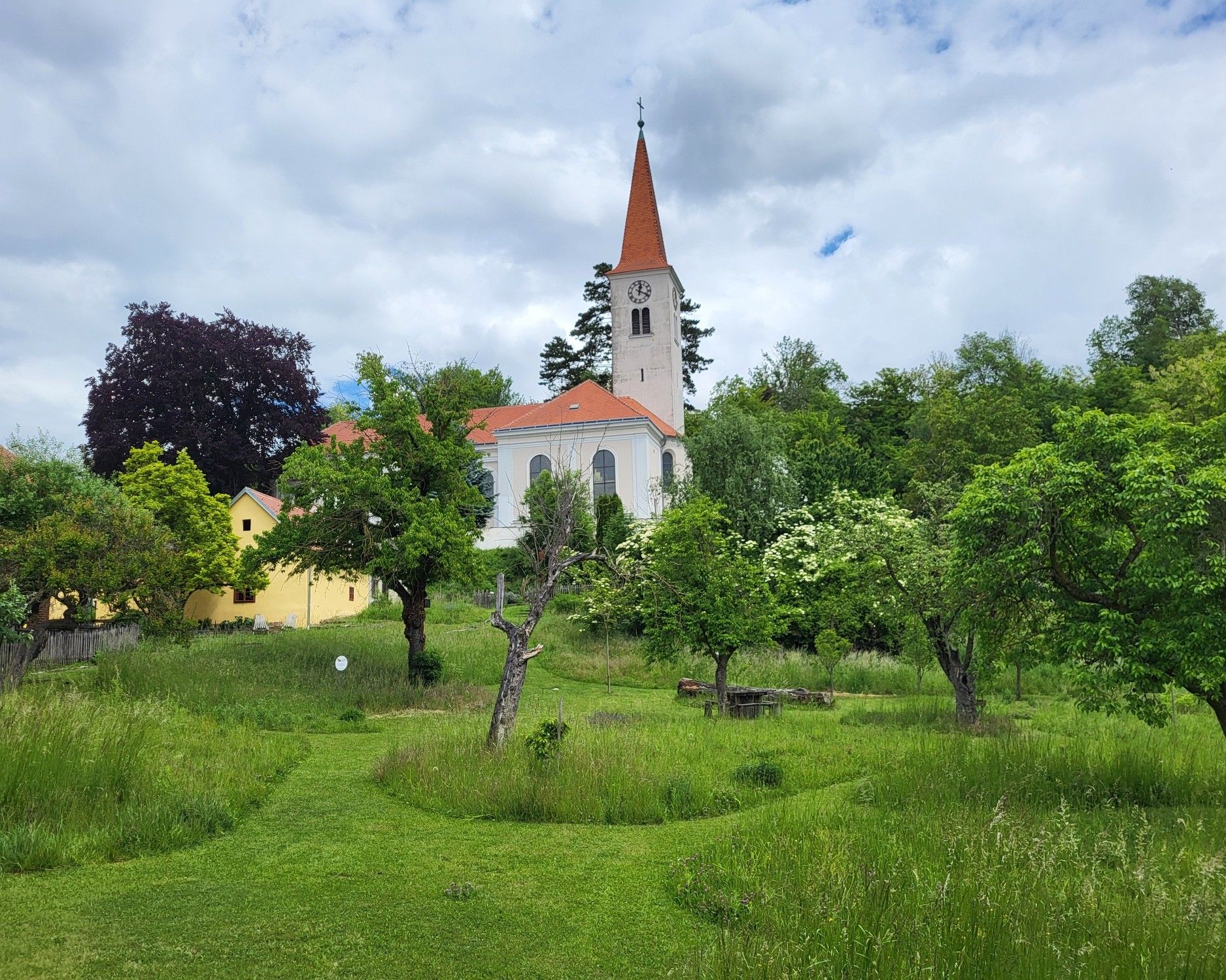 Farská záhrada v Königsbrunne am Wagram s výhľadom na historický kostol, obklopená zelenými lúkami, starými ovocnými stromami a prírodnou záhradou.