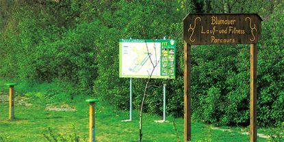 Drevená tabuľa s nápisom "Blumauer Lauf- und Fitness Parcours" pred informačnou tabuľou v prírode.