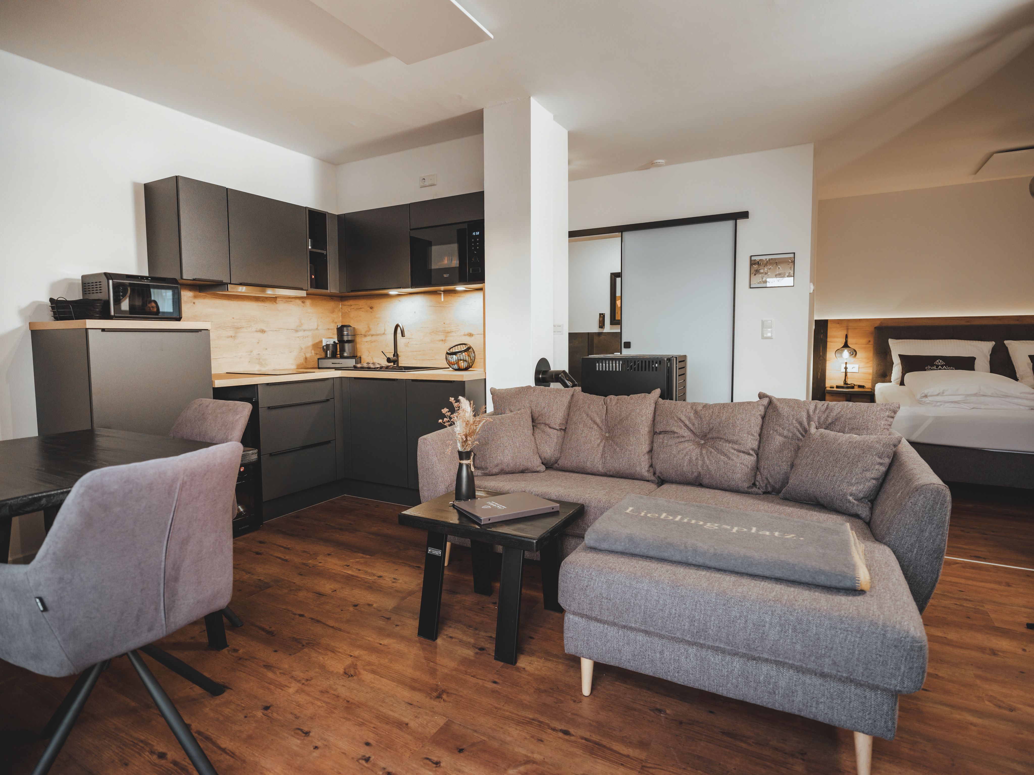 Moderný obývací priestor so sivou pohovkou, kuchynským kútom a jedálenským stolom.