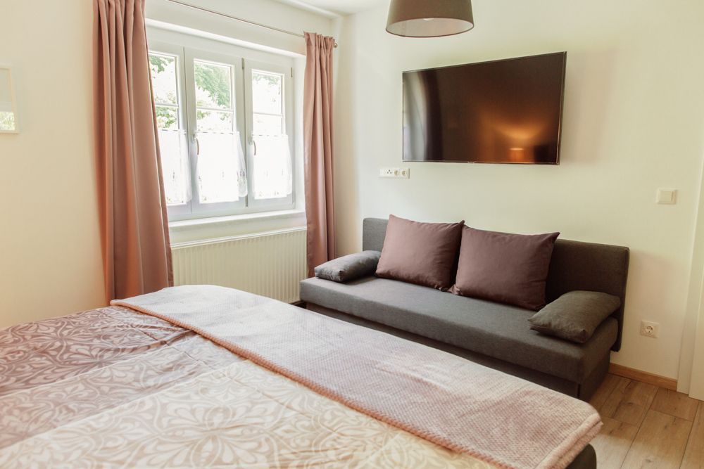 Pohľad do interiéru spálne s posteľou, pohovkou, oknom a nástenným televízorom.