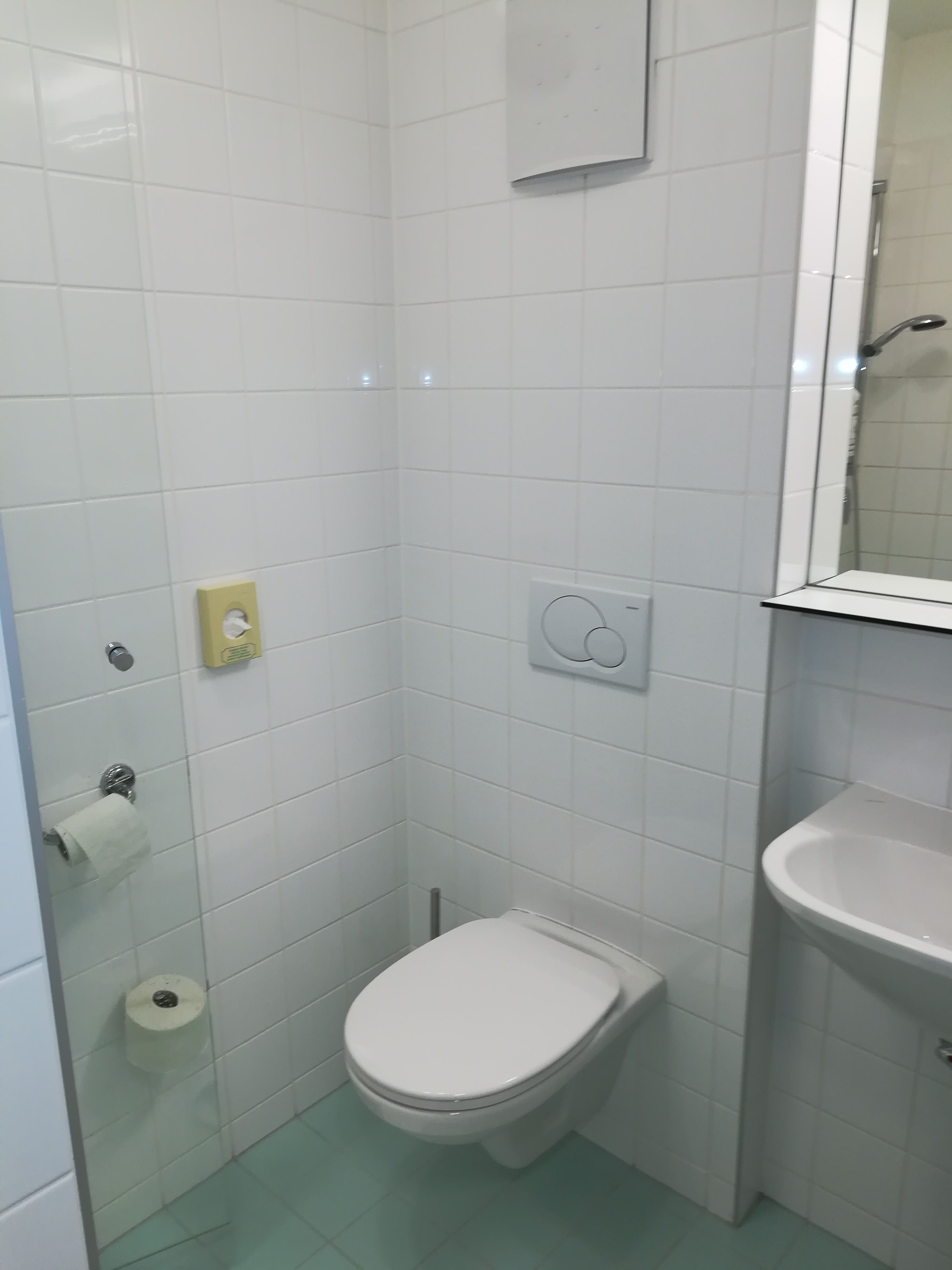 Malá kúpeľňa s bielymi obkladmi, toaletou, umývadlom a držiakom na toaletný papier.