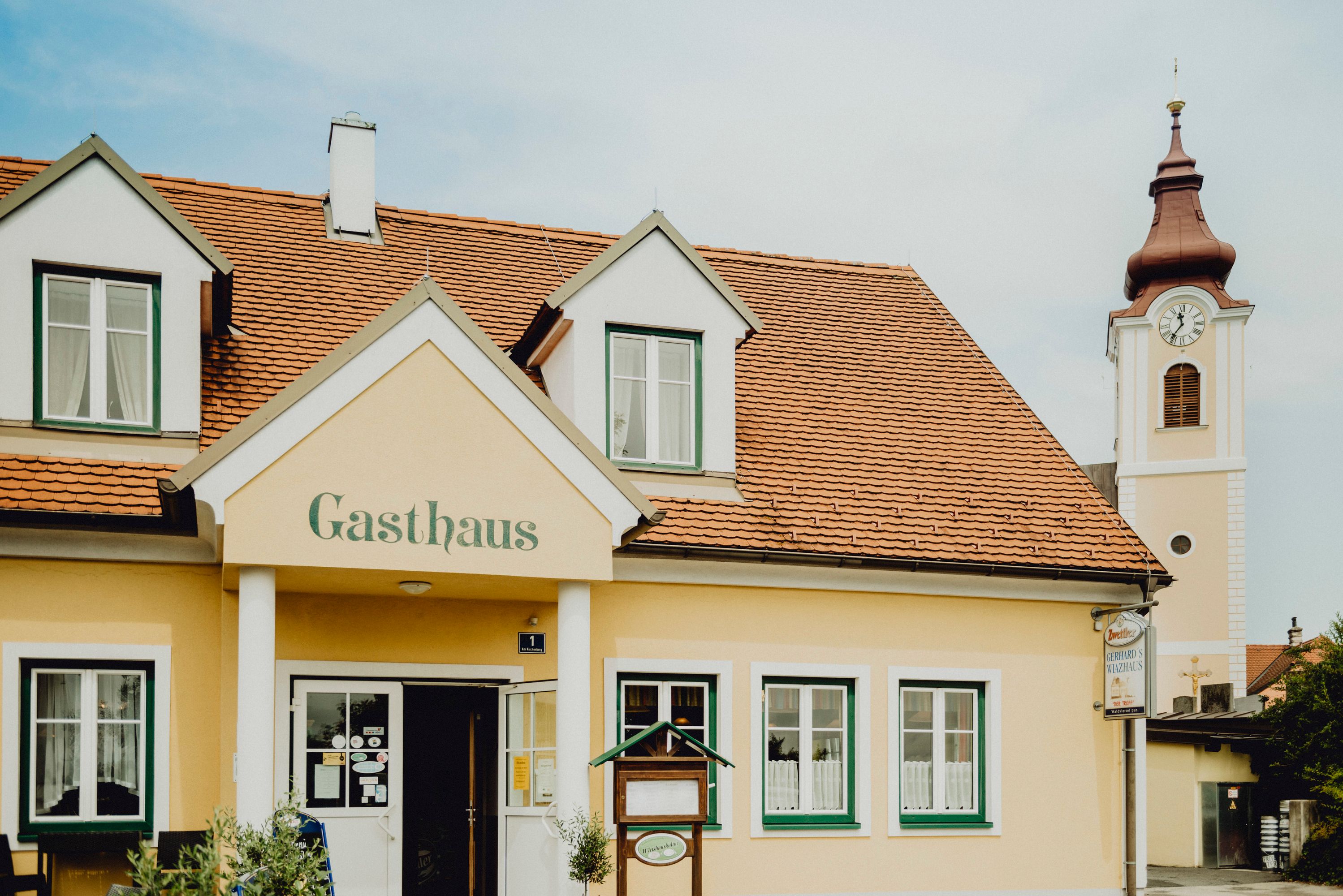 Žltý hostinec s červenými škridlami na streche a kostolnou vežou v pozadí.