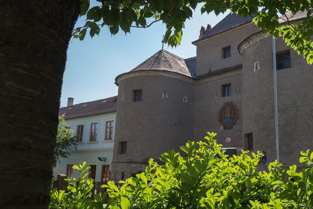 Historická budova s okrúhlymi vežami a erbom, obklopená zelenými stromami a kríkmi.