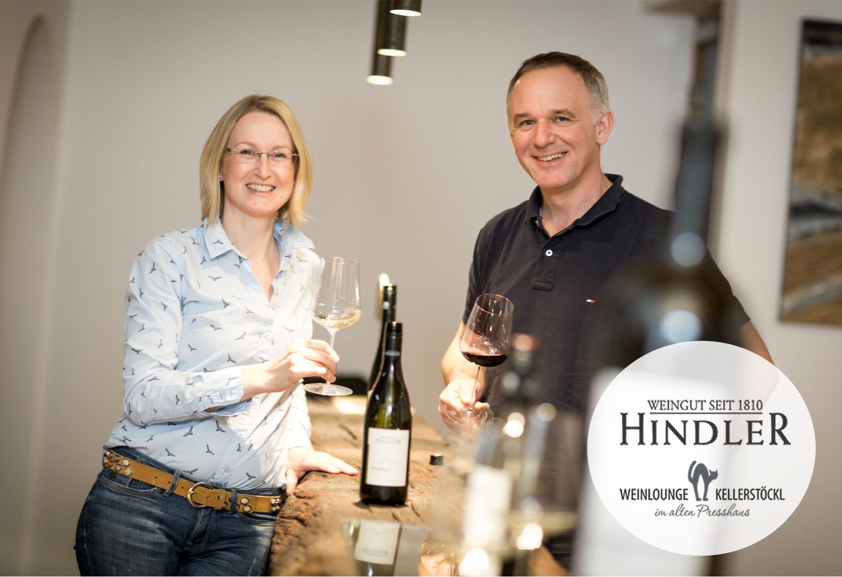 Dvojica vinárov stojí s úsmevom s pohármi na víno v rukách pri stole vo vinárstve Hindler.