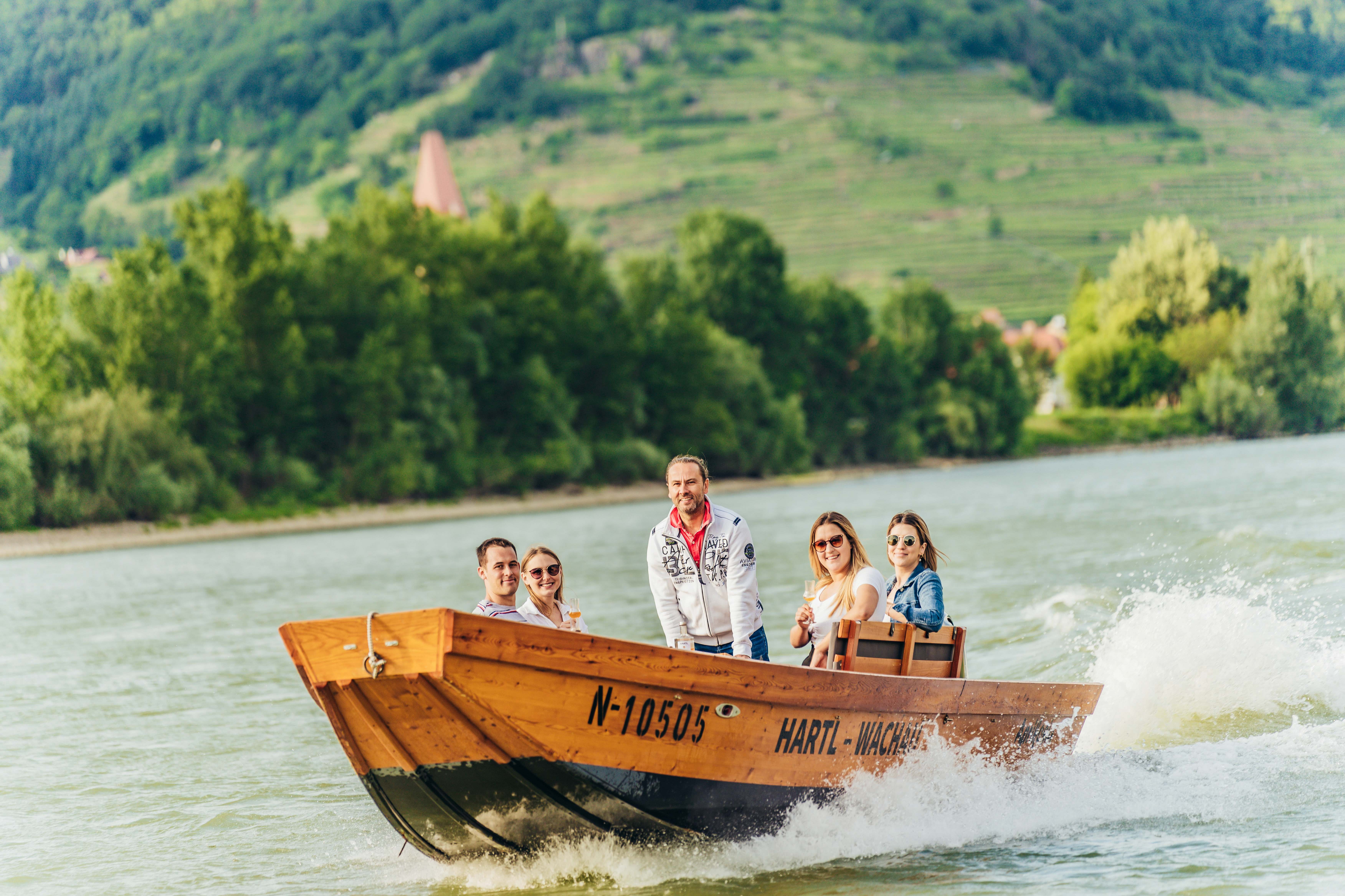 Skupina ľudí na drevenej lodi na Dunaji so zelenou krajinou v pozadí.