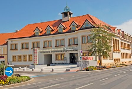 Radnica v Traiskirchene s červenou strechou a niekoľkými oknami, obklopená ulicou a kvetinovými záhonmi.
