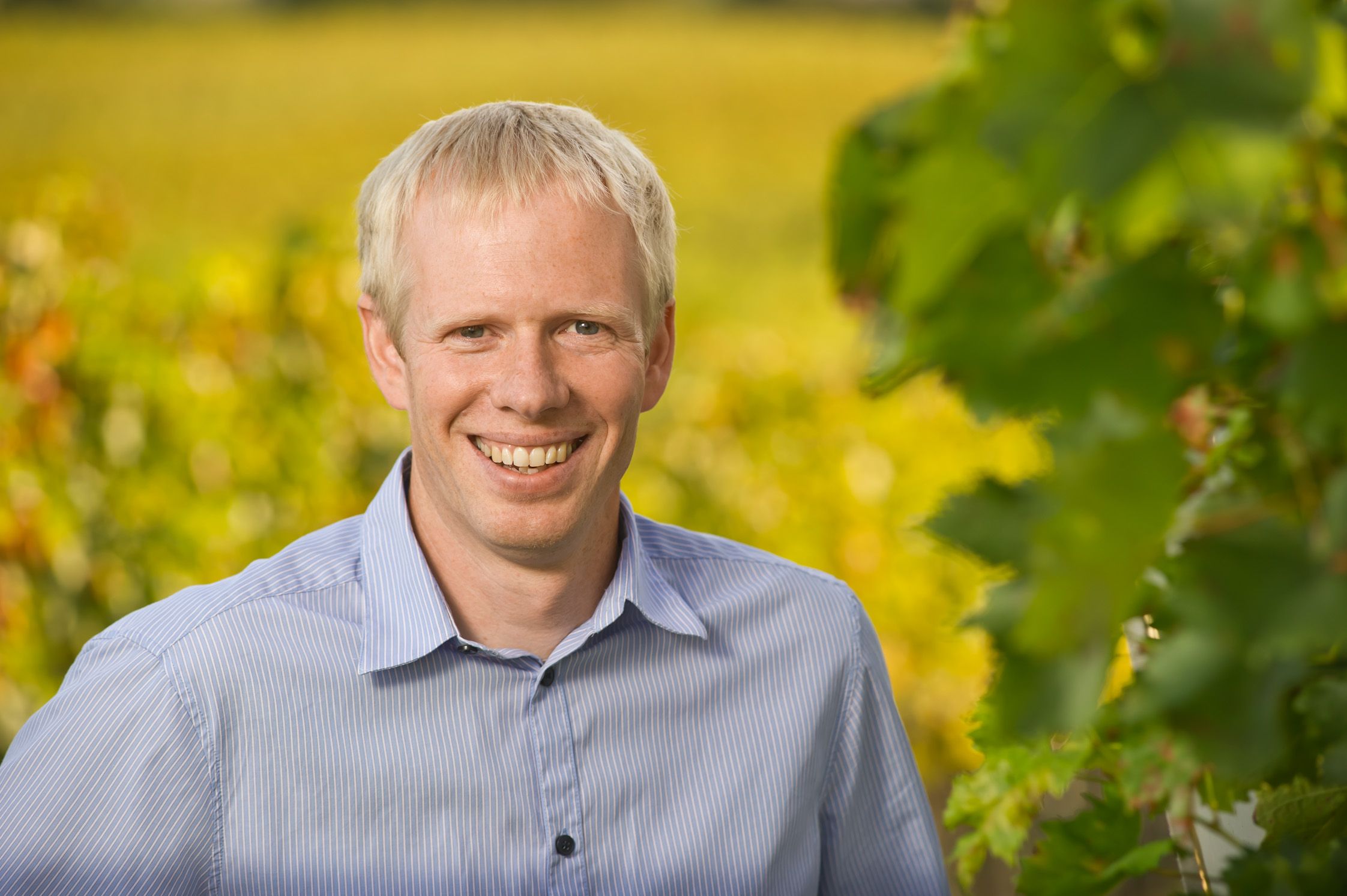 Usmievajúci sa muž v modrej košeli stojí pred rozmazaným pozadím so zelenými listami.