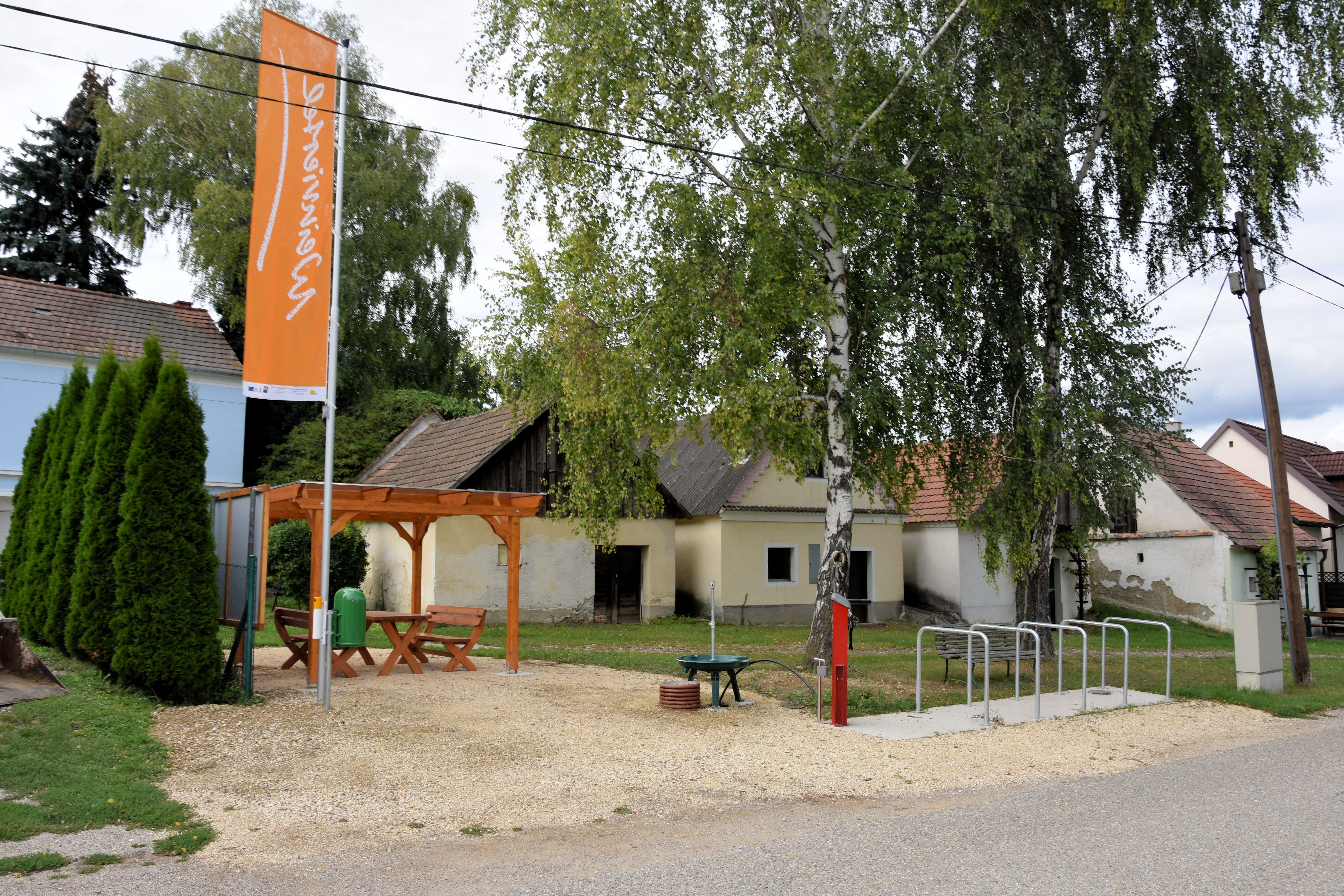 Odpočívadlo s dreveným stolom a lavicami pod altánkom, obklopené stromami a starými budovami. K dispozícii je aj stojan na bicykle a informačná tabuľa.