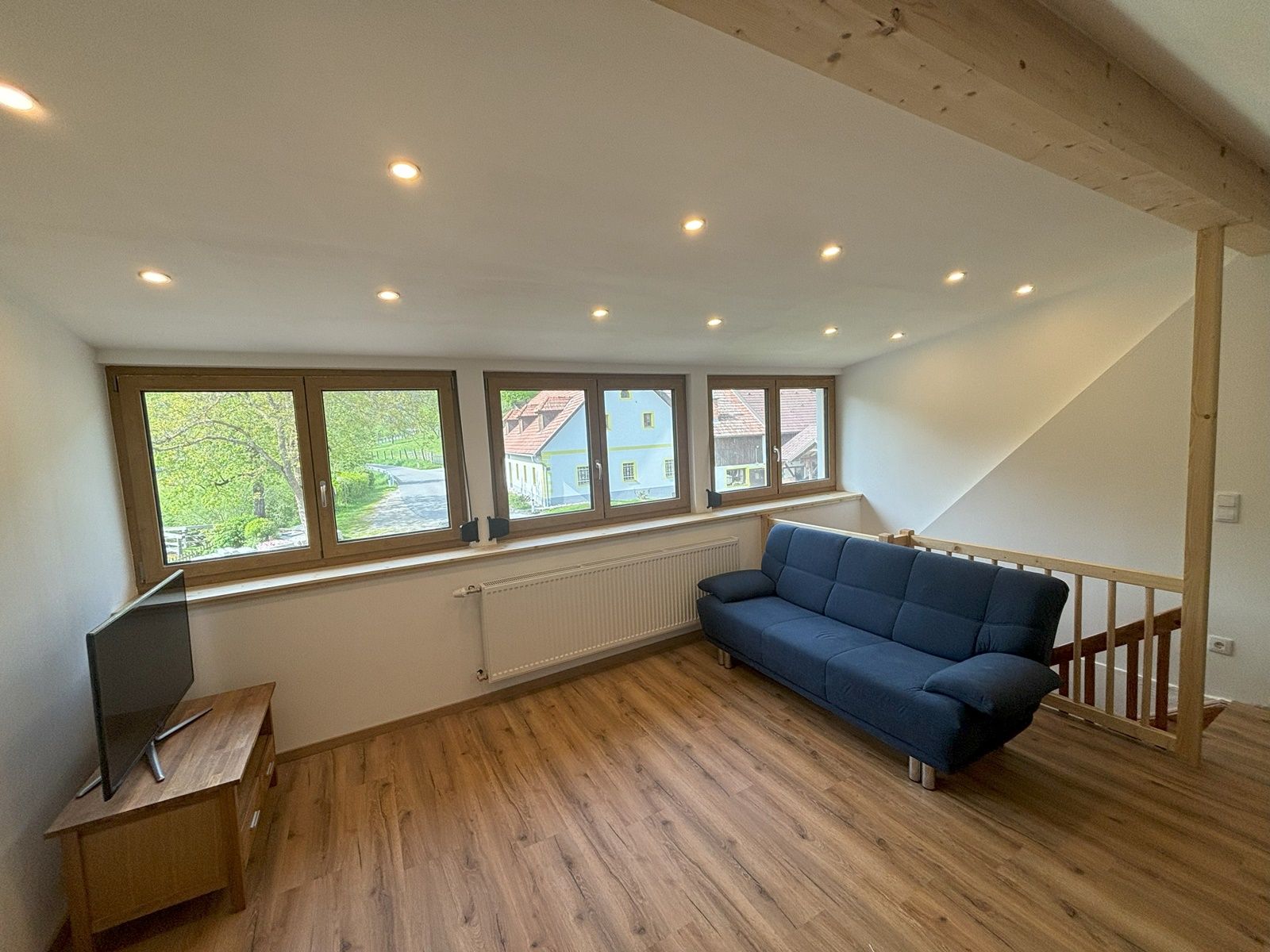 Obývacia izba s modrou pohovkou, drevenou podlahou a veľkými oknami s výhľadom von.