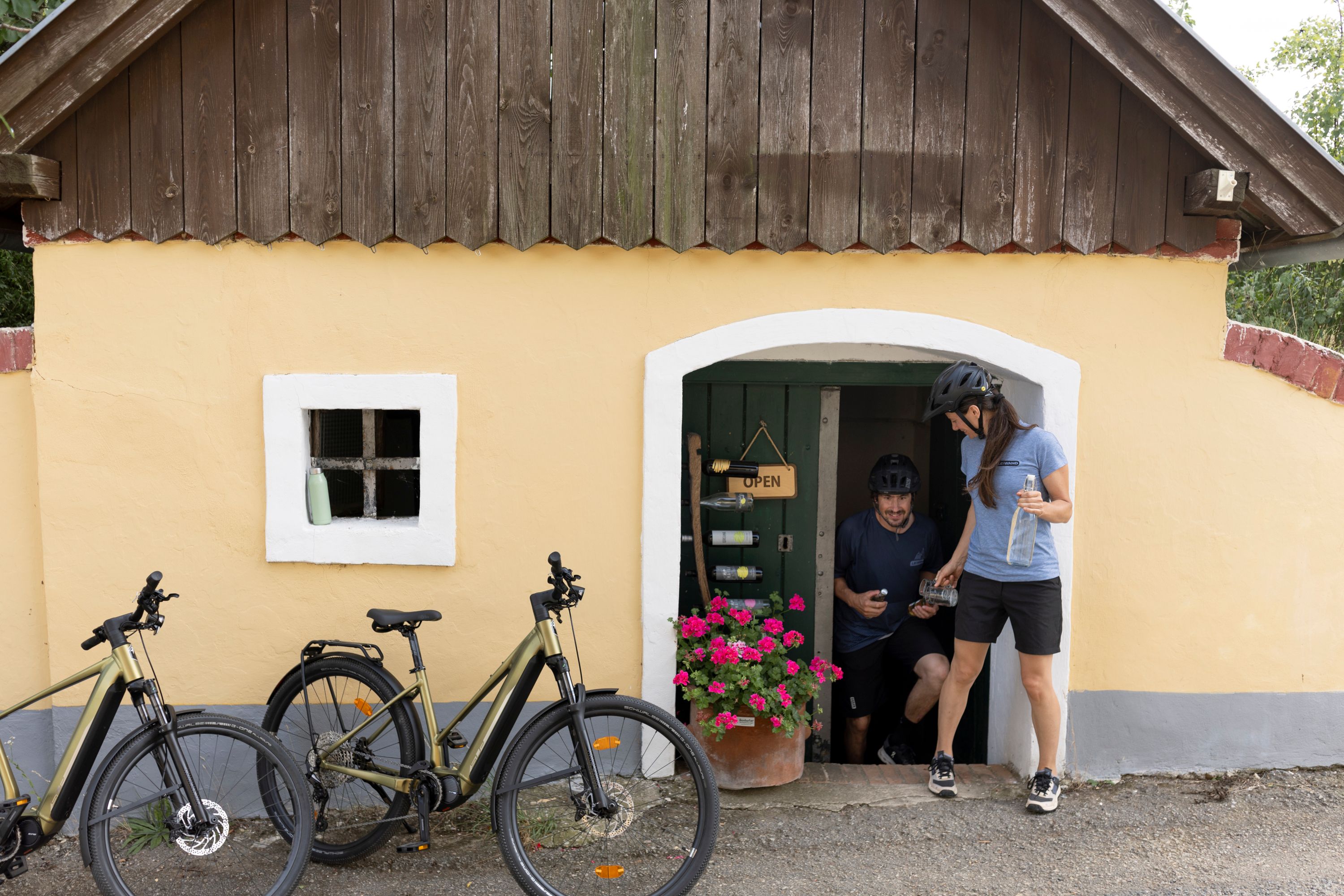 Dvaja ľudia s bicyklami pred malou žltou budovou s drevenou strechou a kvetmi.