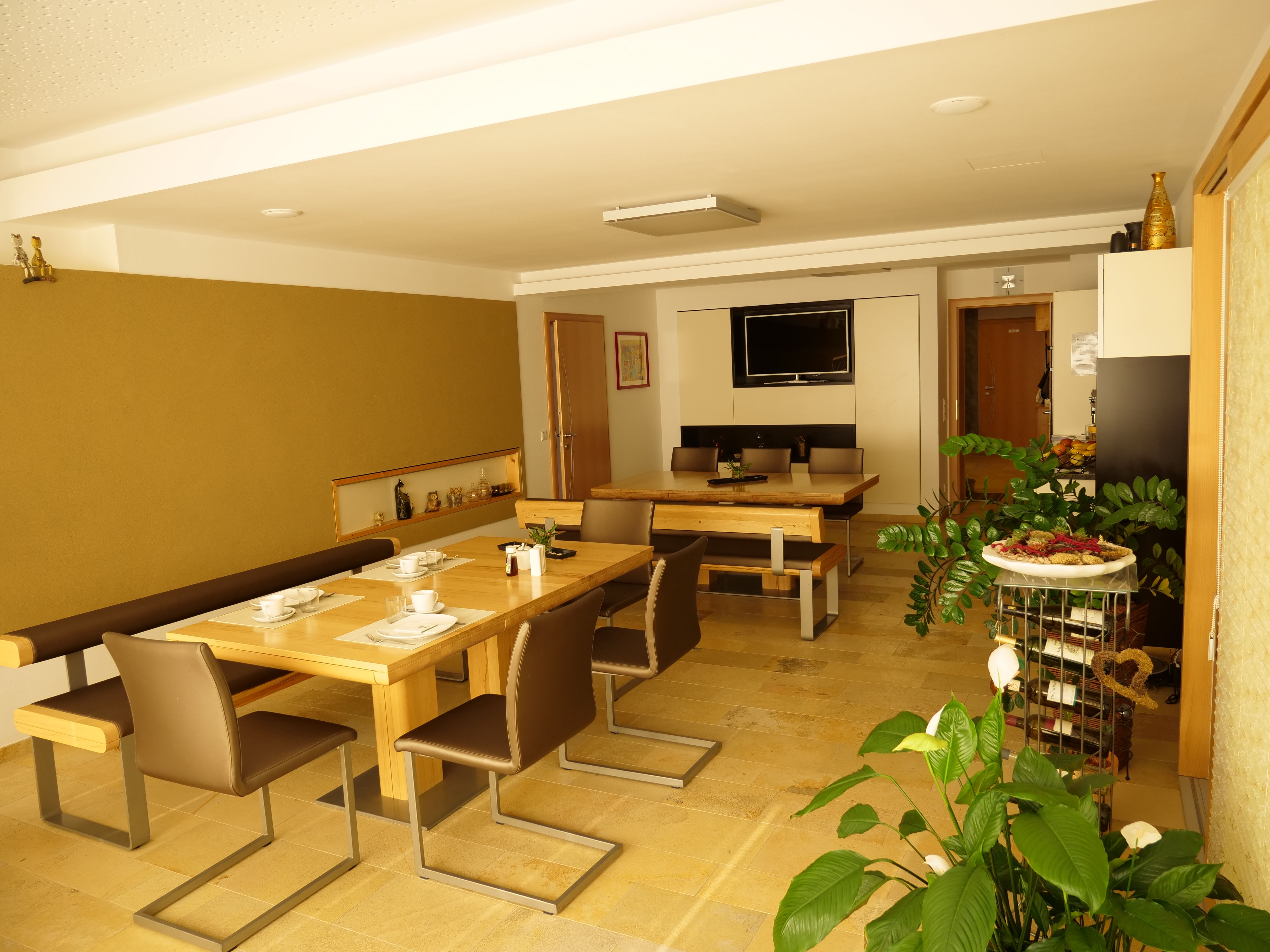 Raňajková miestnosť s drevenými stolmi, stoličkami a rastlinami, zdobená riadom a televízorom na stene.