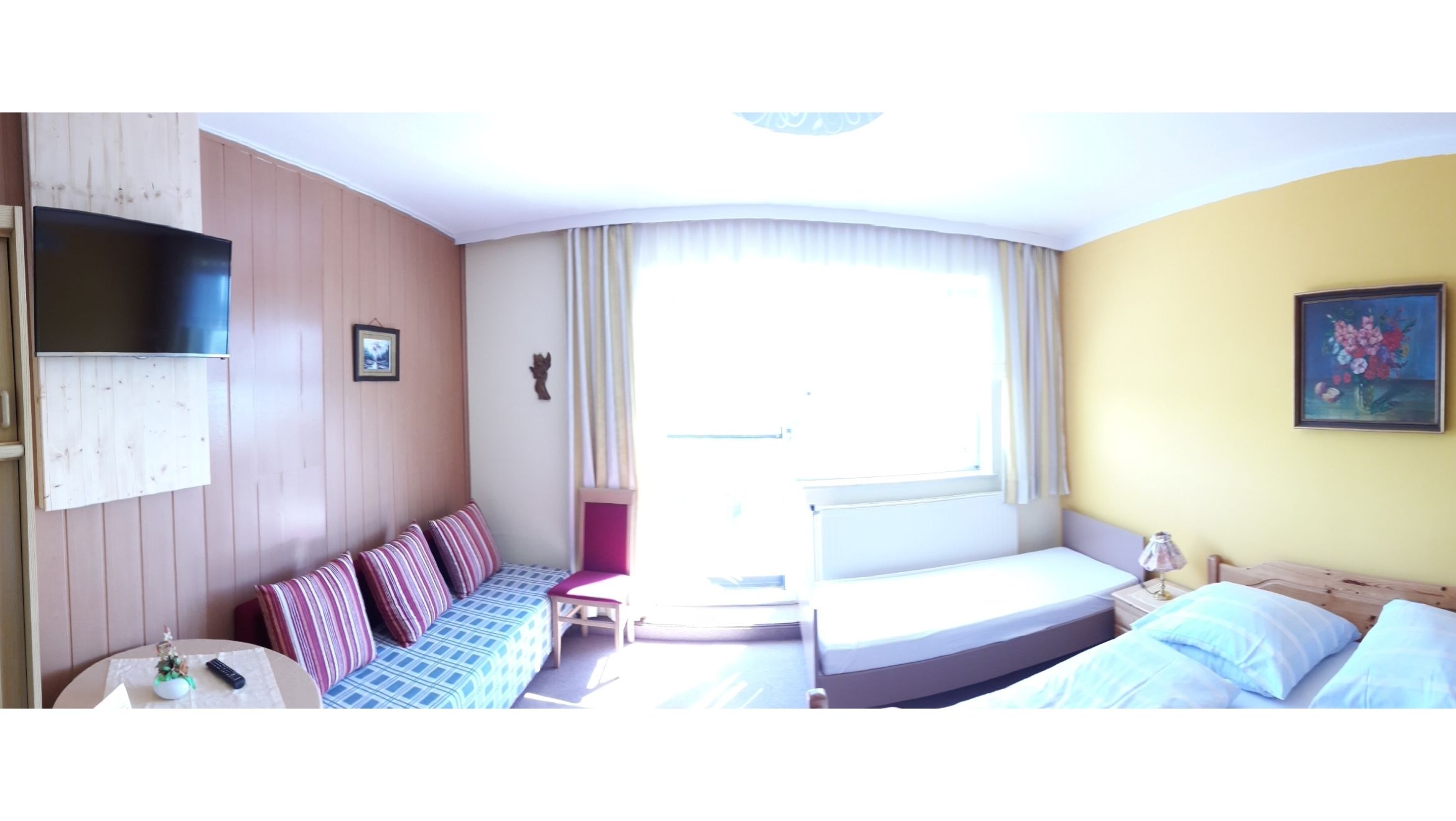 Svetlá izba pre hostí s pohovkou, posteľou, TV a maľbou na stene.