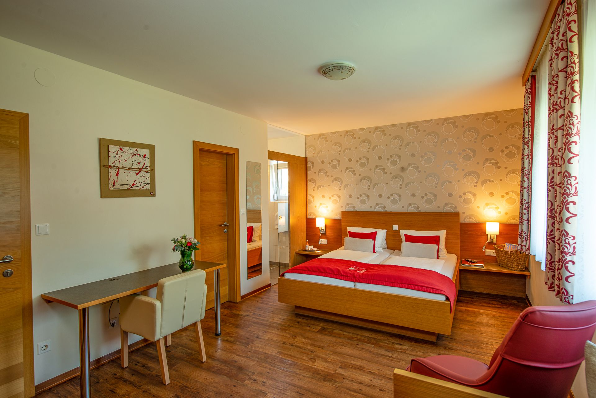 Hotelová izba s manželskou posteľou, písacím stolom, stoličkou a červeným kreslom.