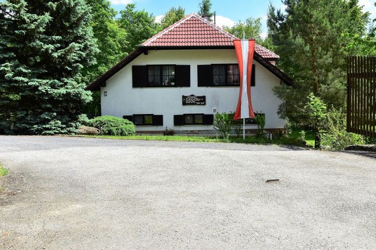 Biely dom s červenou strechou a rakúskou vlajkou pred ním, obklopený stromami.