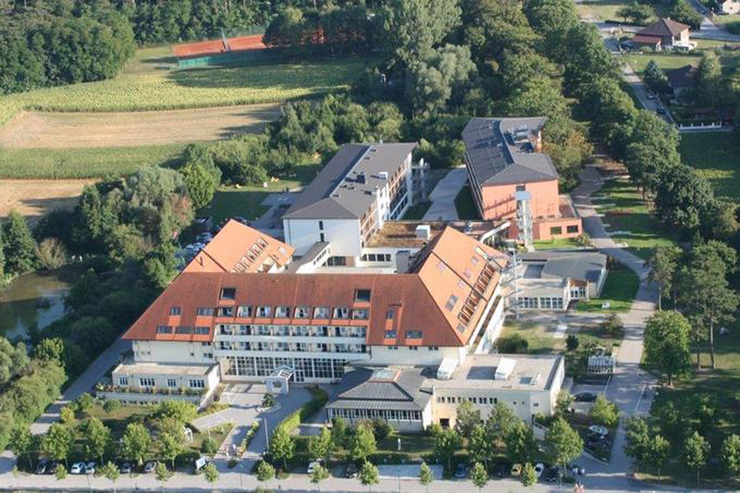 Letecký pohľad na kliniku s červenými strechami, obklopenú stromami a poľami.