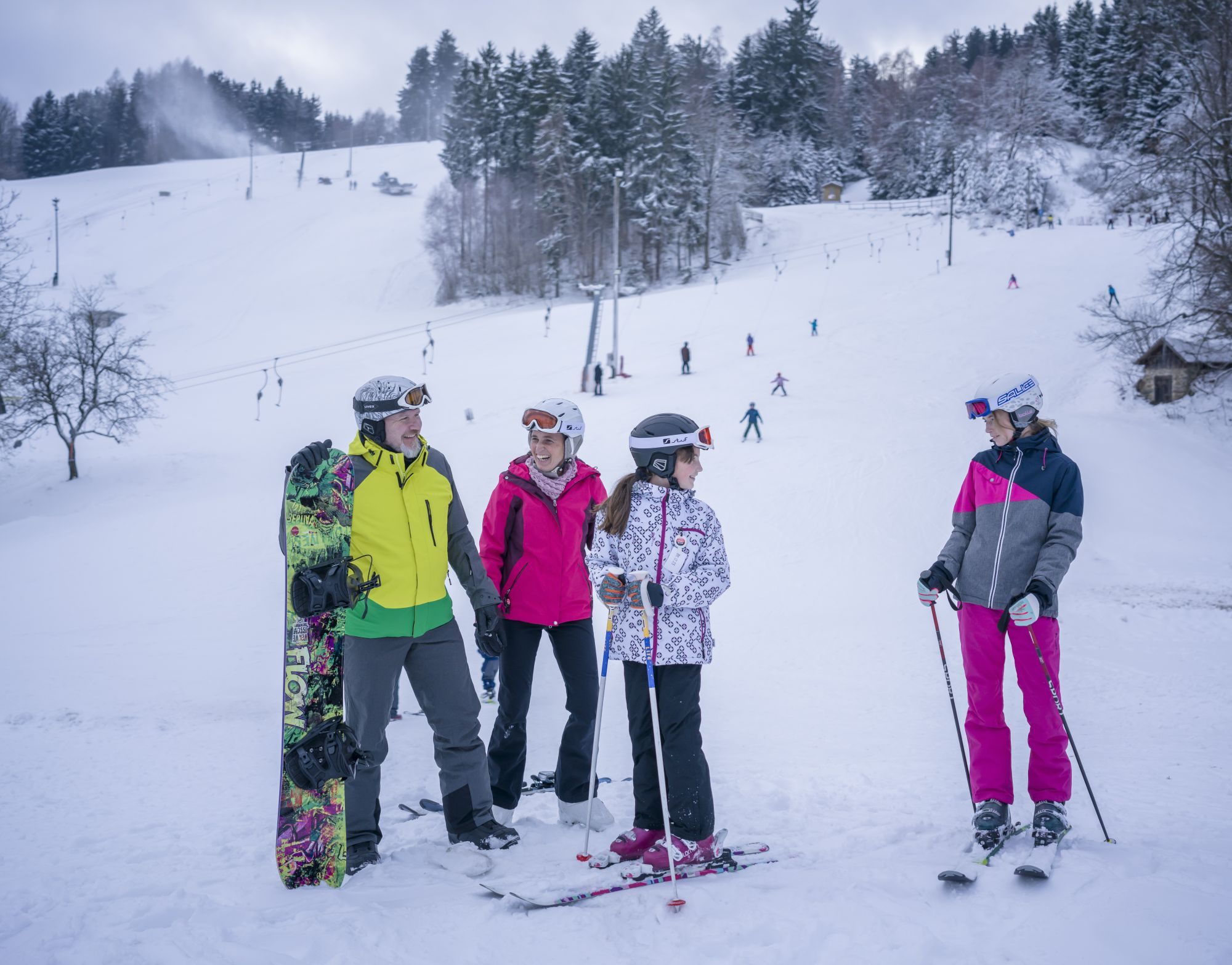 Skupina lyžiarov a snowboardistov na snehu pred lyžiarskym svahom s vlekom.