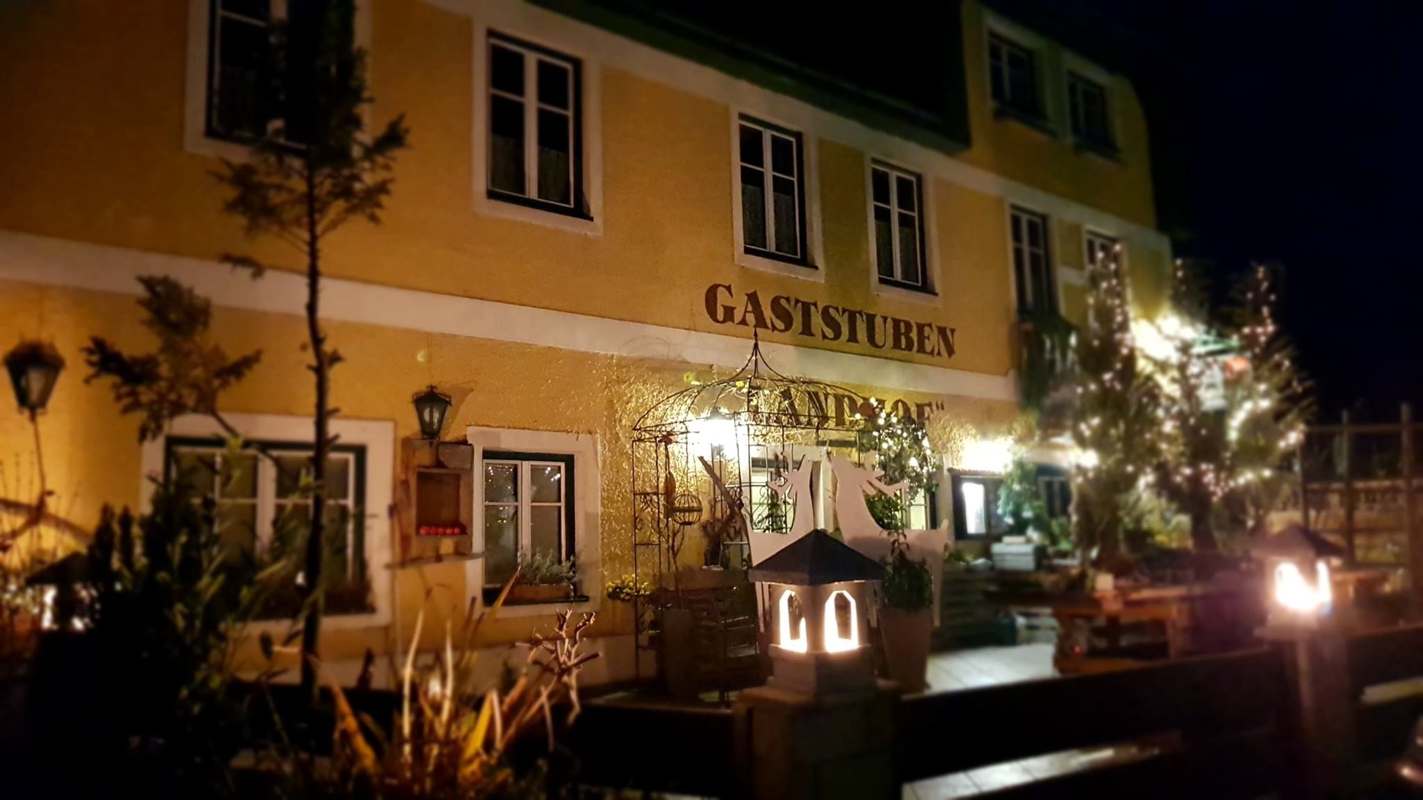 Nočný pohľad na žltú budovu s nápisom "Gaststuben Landhof", osvetlenú lampášmi a obklopenú rastlinami.