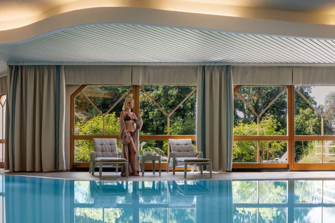 Žena v plavkách stojí pri krytom bazéne s veľkými oknami s výhľadom na zelené stromy.