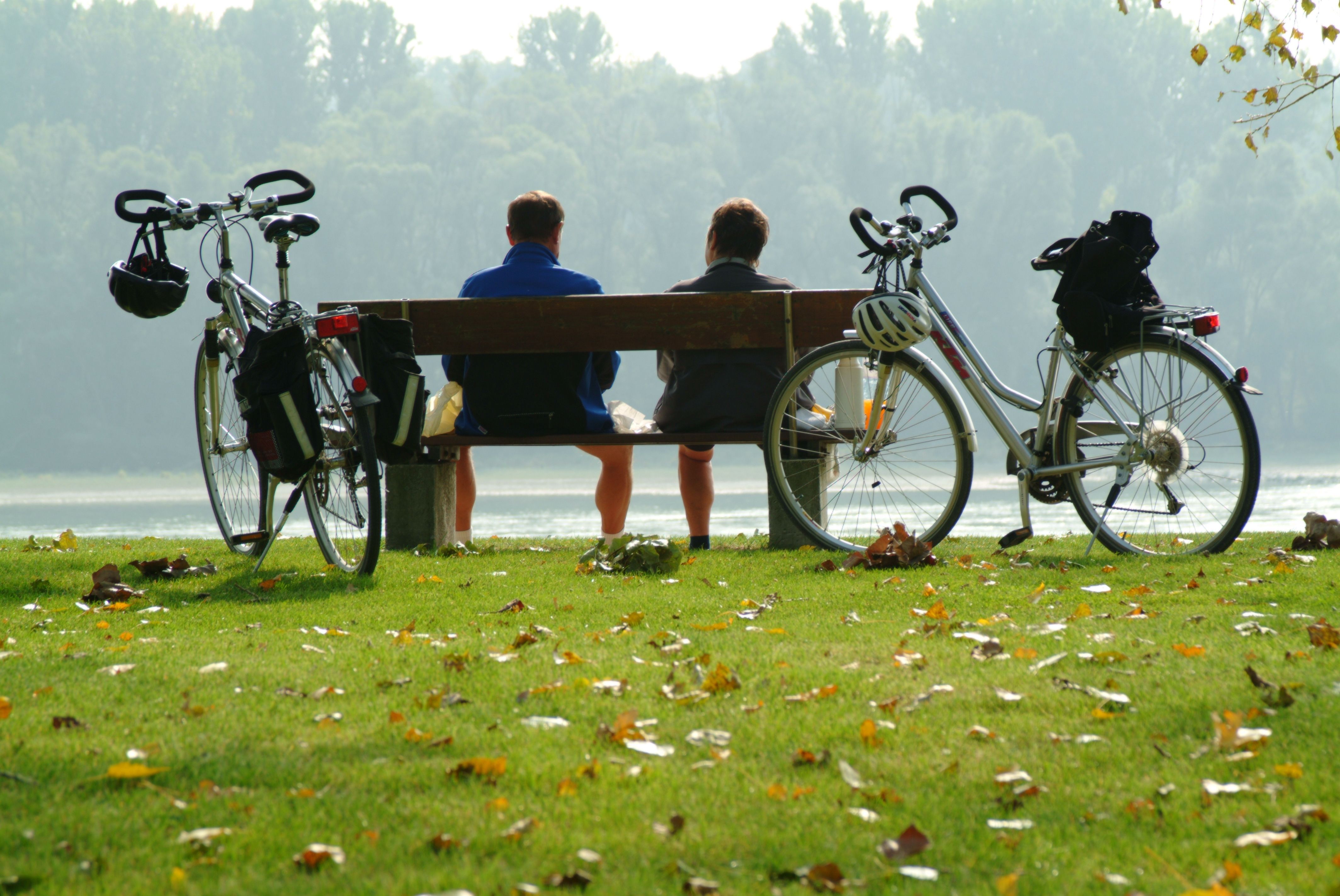 Dvaja ľudia sedia na lavičke pri jazere, vedľa nich sú bicykle, obklopené jesenným lístím.