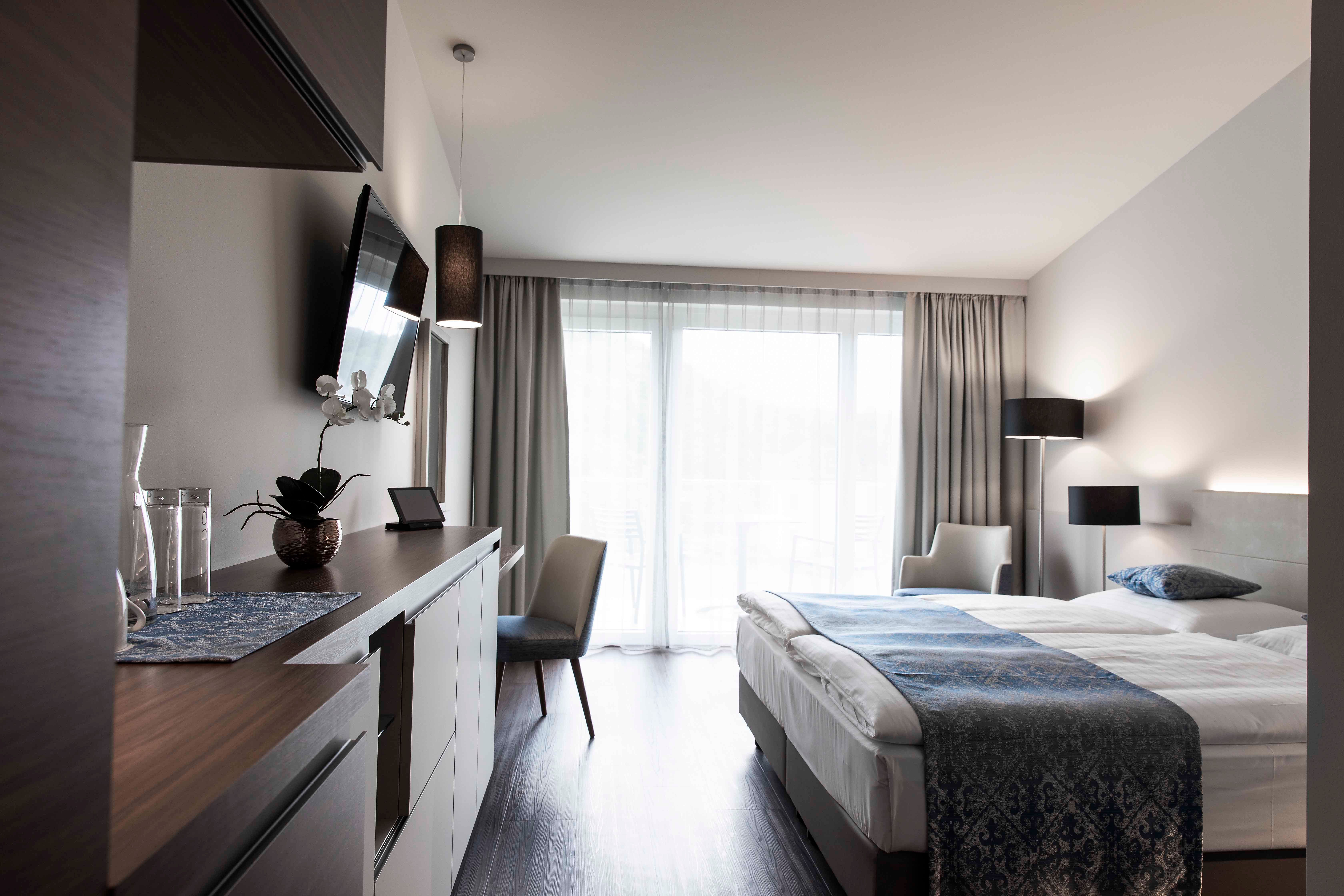 Moderná hotelová izba s manželskou posteľou, TV, písacím stolom a veľkými oknami so závesmi.