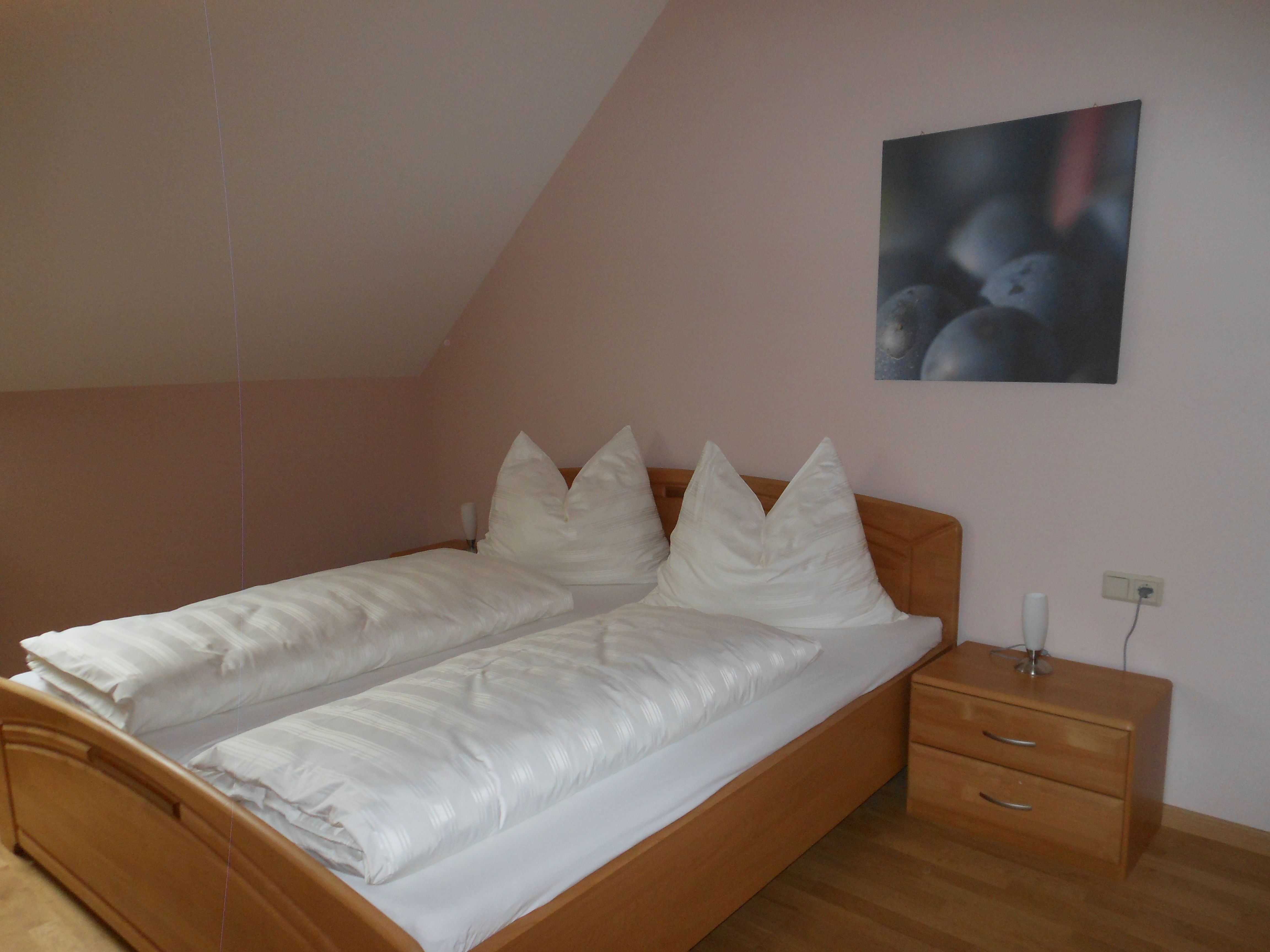 Jednoduchá izba pre hostí s manželskou posteľou, bielymi perinami a nočným stolíkom s lampou.