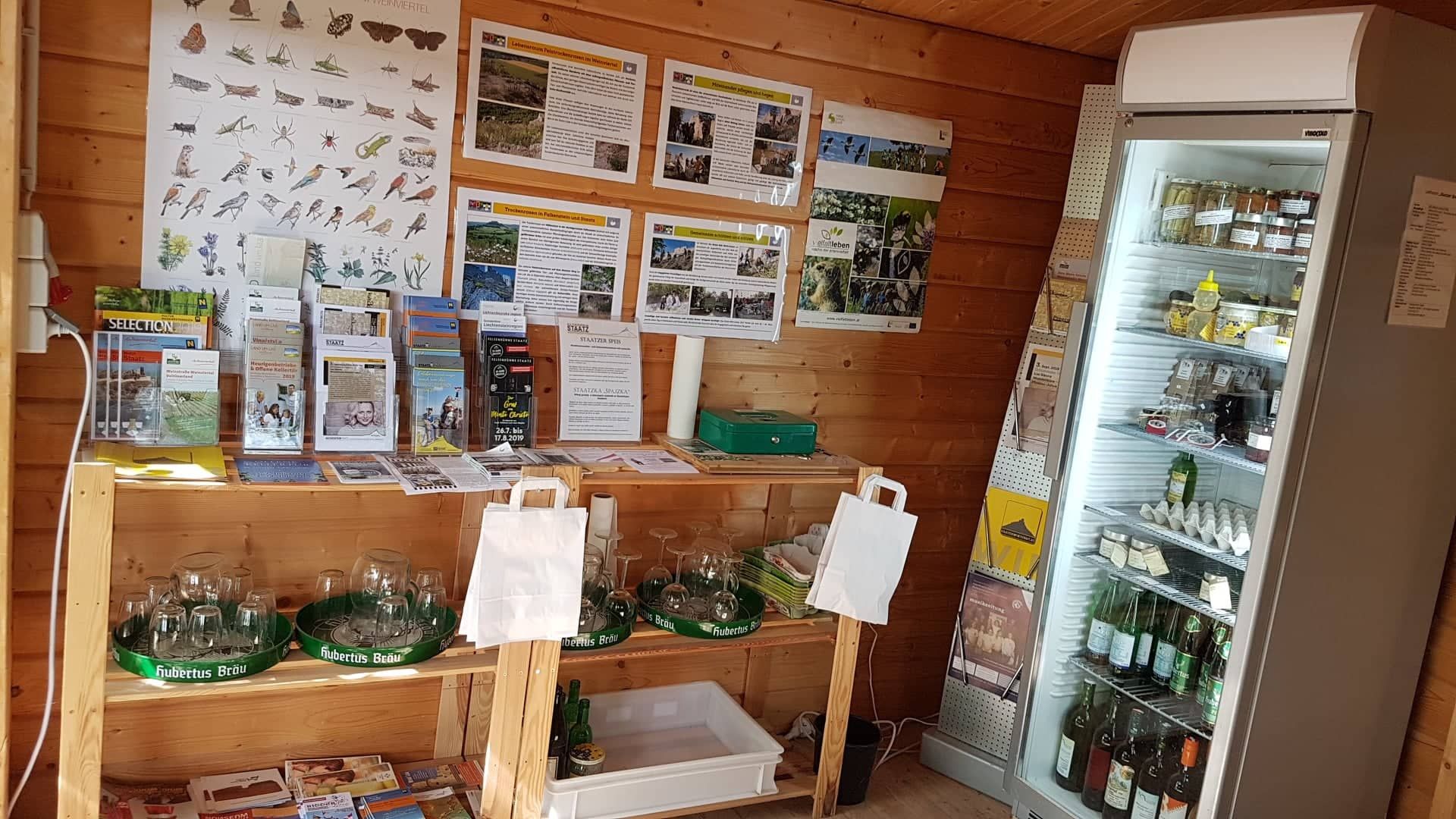 Pohľad do interiéru malého predajného stánku s informačnými materiálmi a chladničkou.