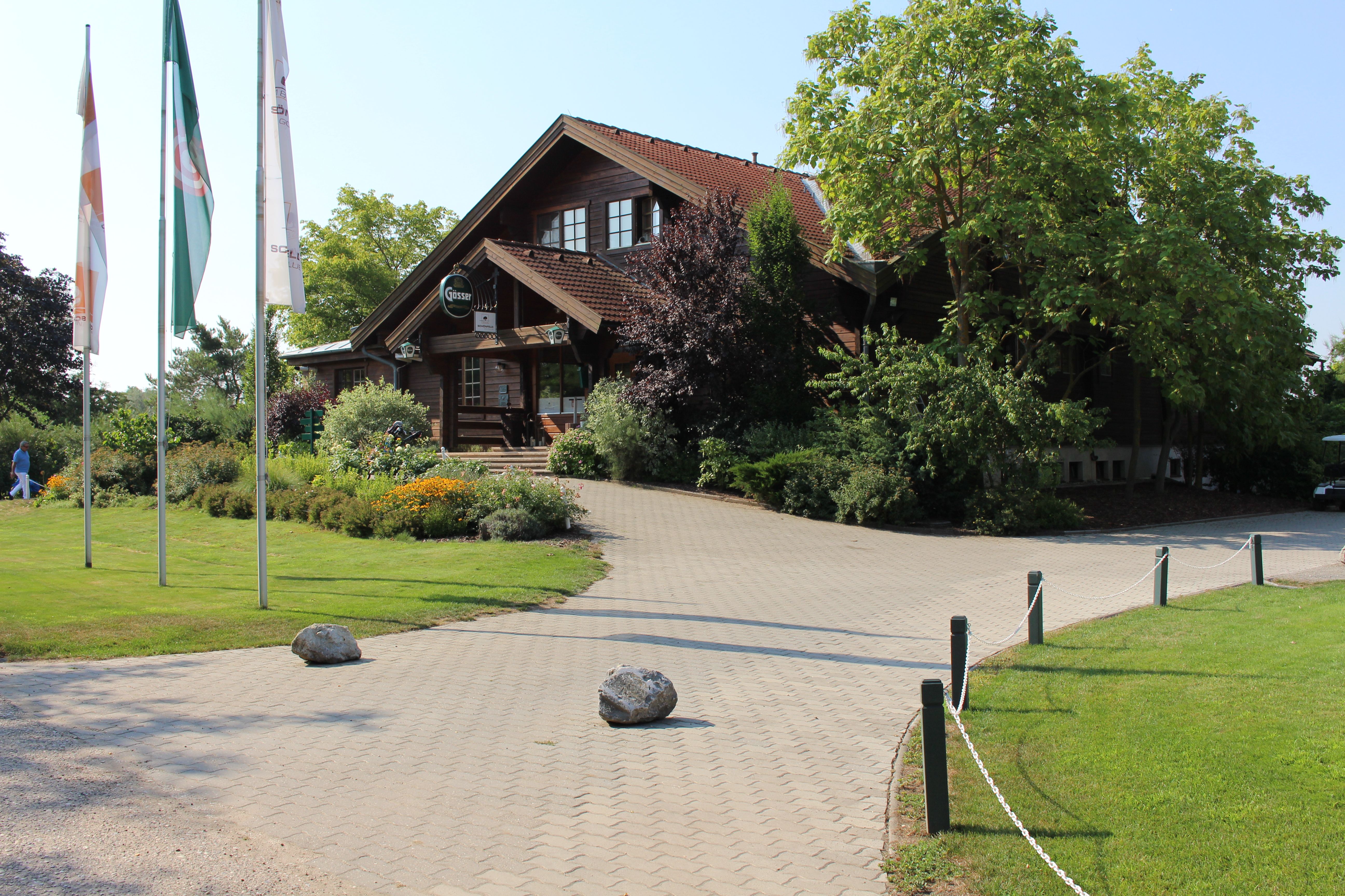 Vchod do golfového klubu s drevenou budovou a vlajkami.