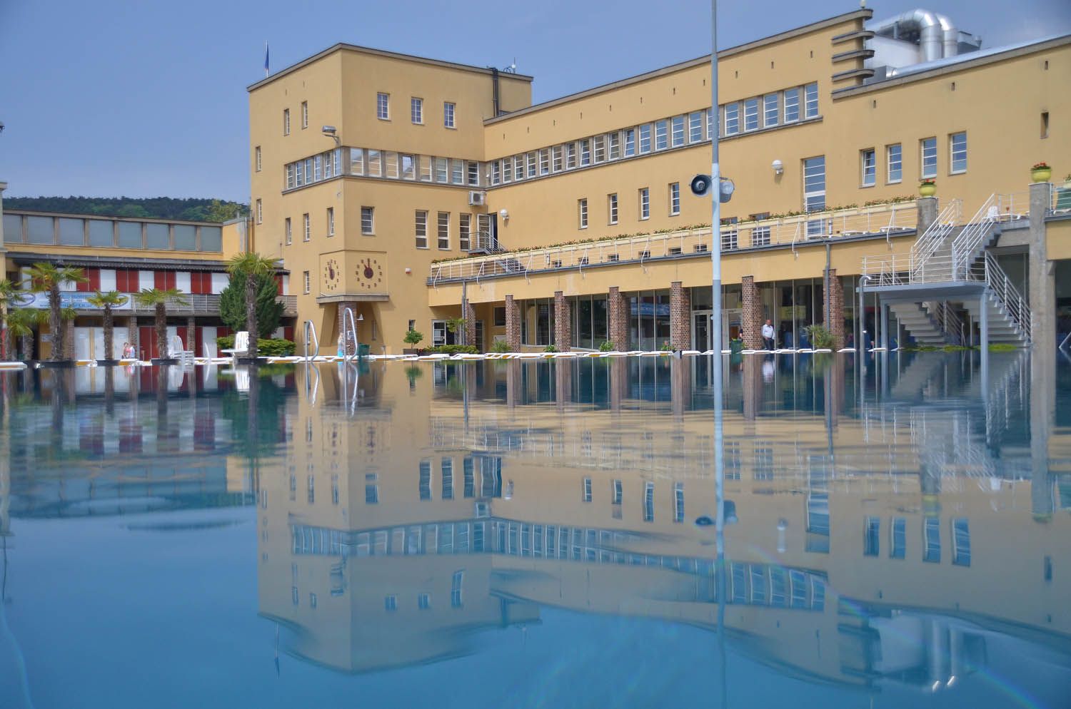 Vonkajší bazén s veľkou žltou budovou v pozadí, ktorá sa odráža vo vode.