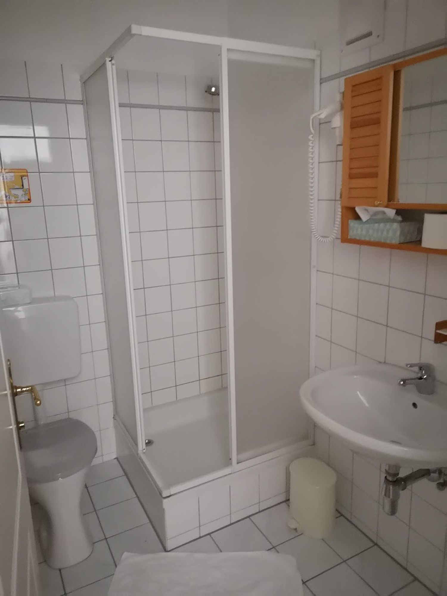 Malá, bielo obložená kúpeľňa so sprchovacím kútom, toaletou a umývadlom.