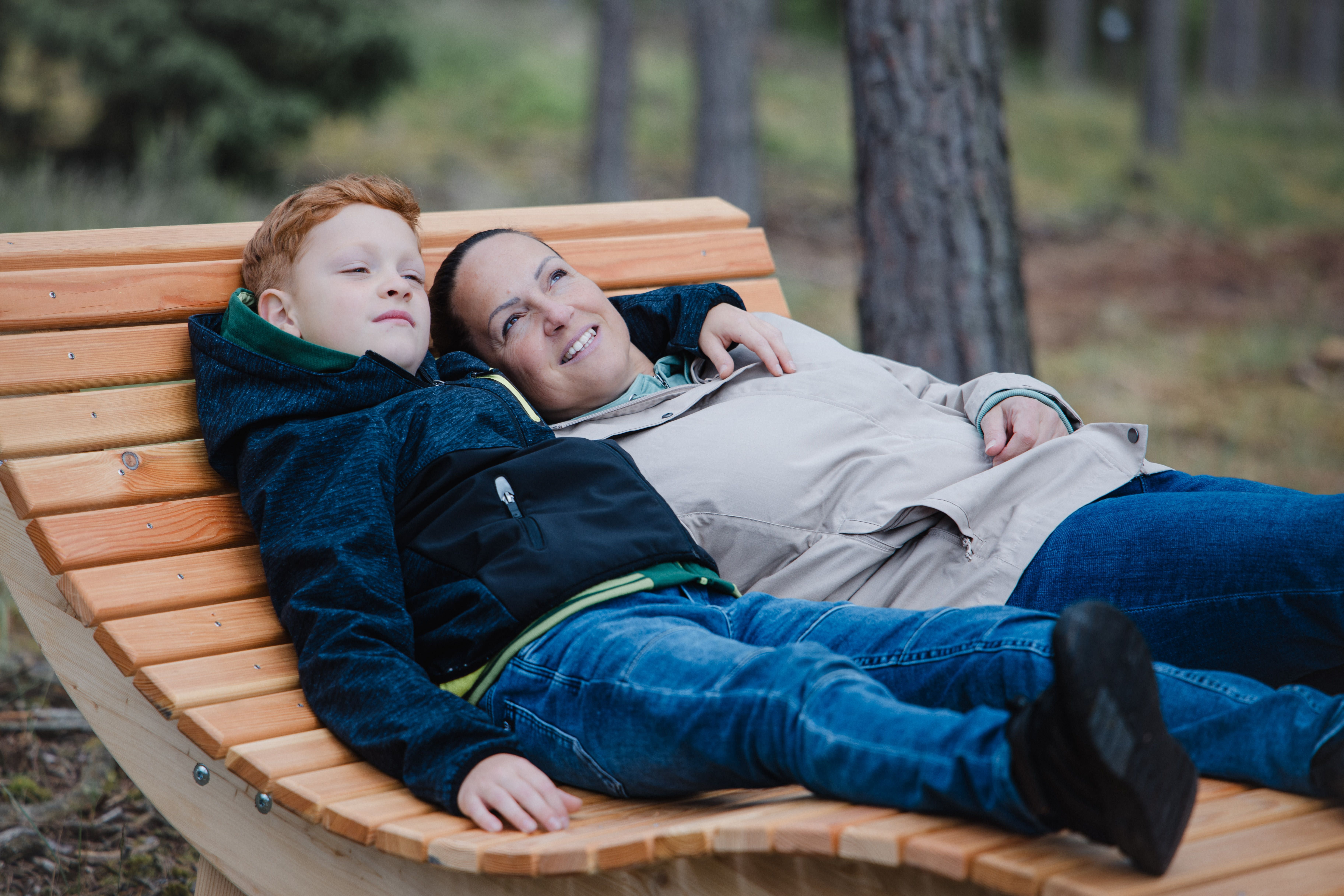 Dvaja ľudia ležia uvoľnene na drevenej lavičke v lese.
