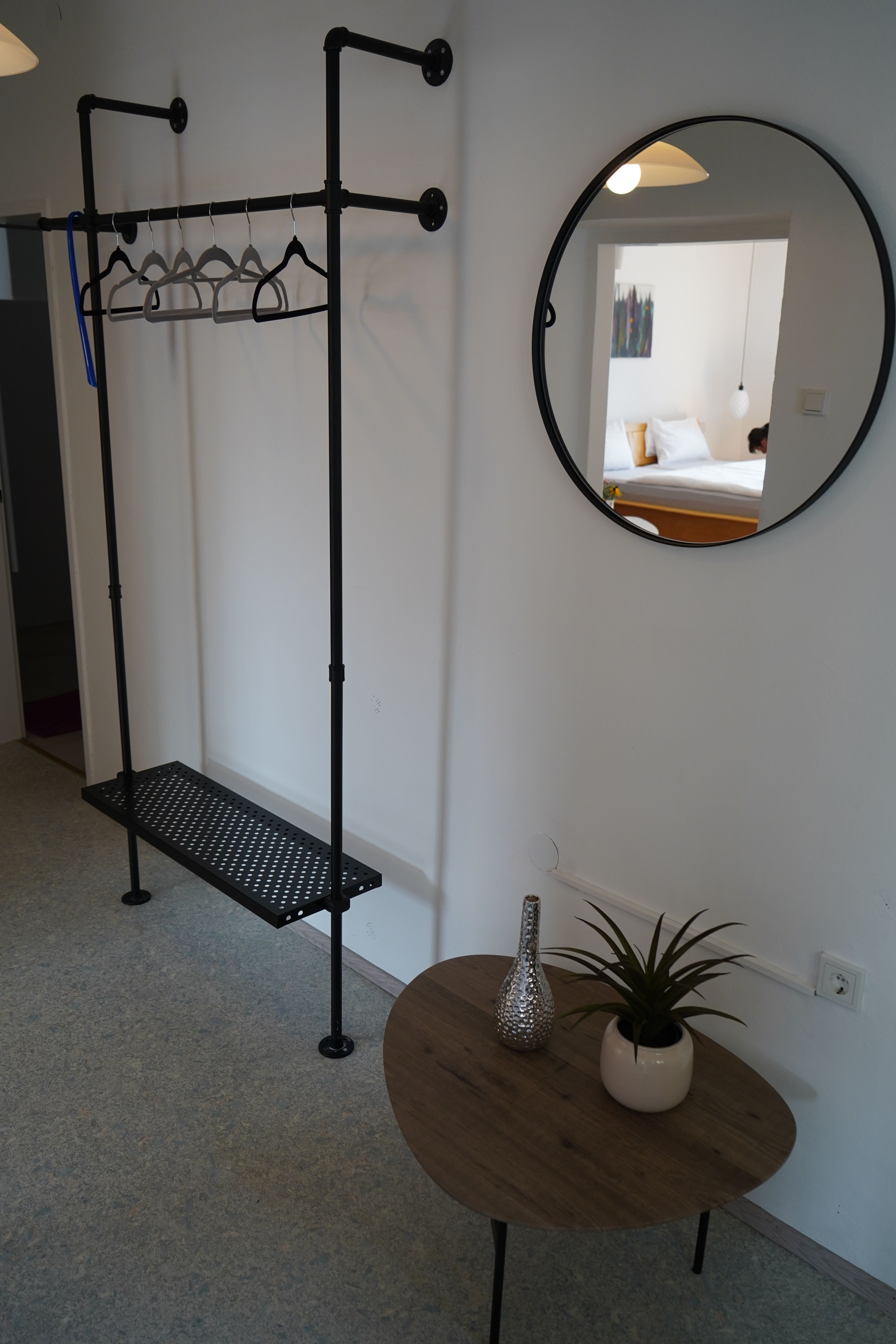 Pohľad do interiéru izby s vešiakom na šaty, okrúhlym zrkadlom a malým stolíkom s rastlinou a vázou.