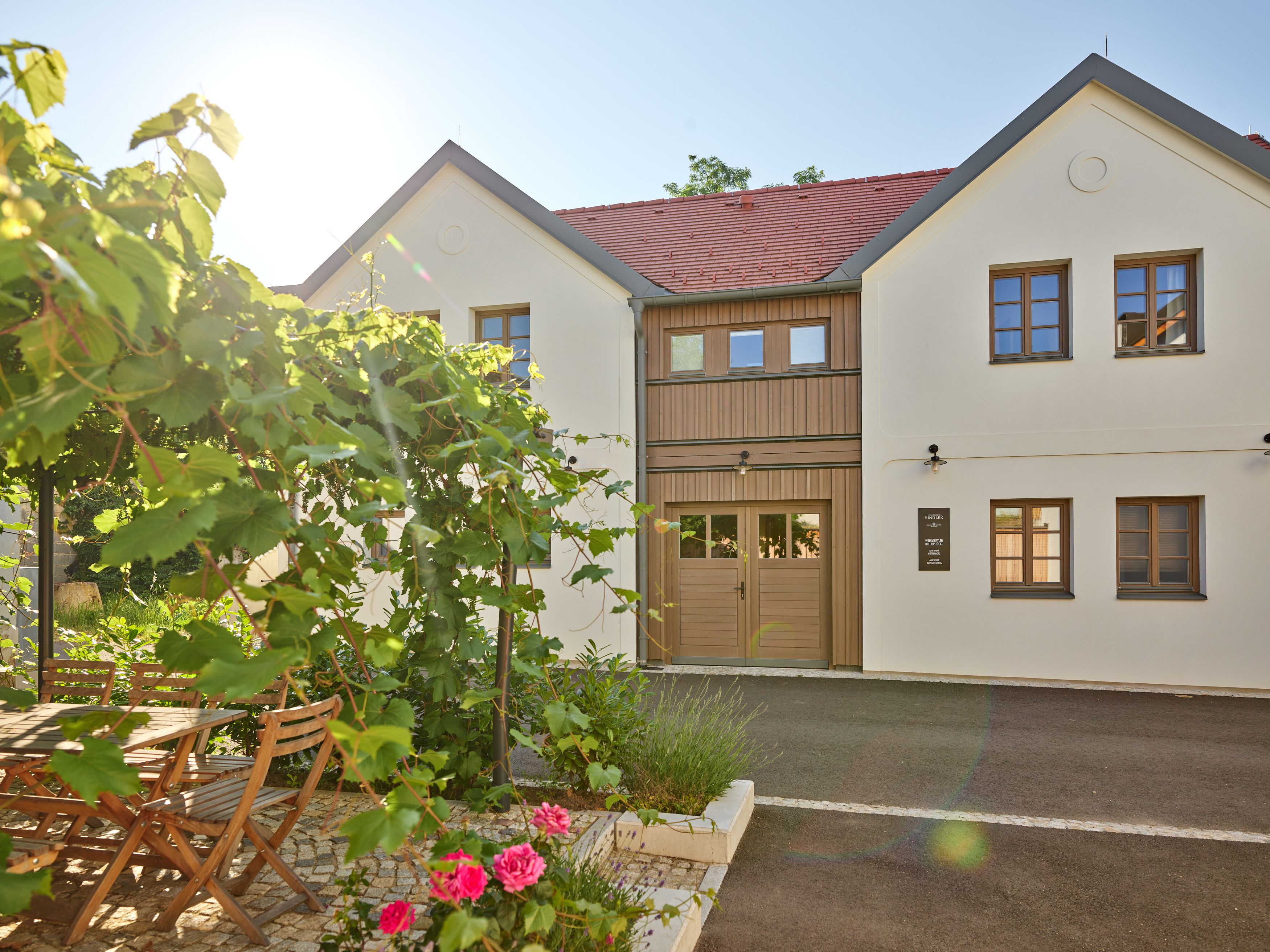 Moderná dvojpodlažná budova s bielymi stenami a červenými škridlami na streche. V popredí záhrada s dreveným stolom, stoličkami a kvitnúcimi rastlinami.