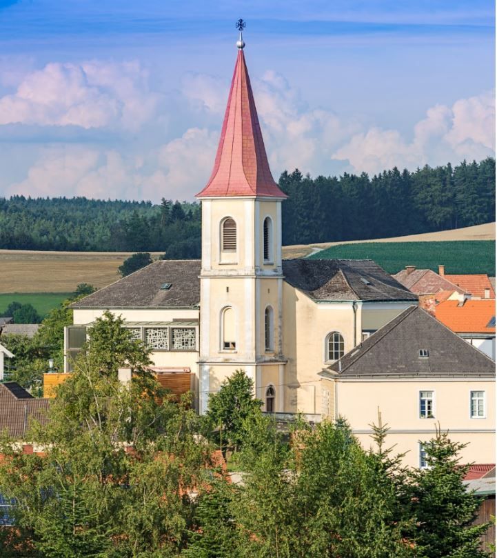 Farský kostol Hochwolkersdorf s červenou špicatou strechou veže a okolitou krajinou.