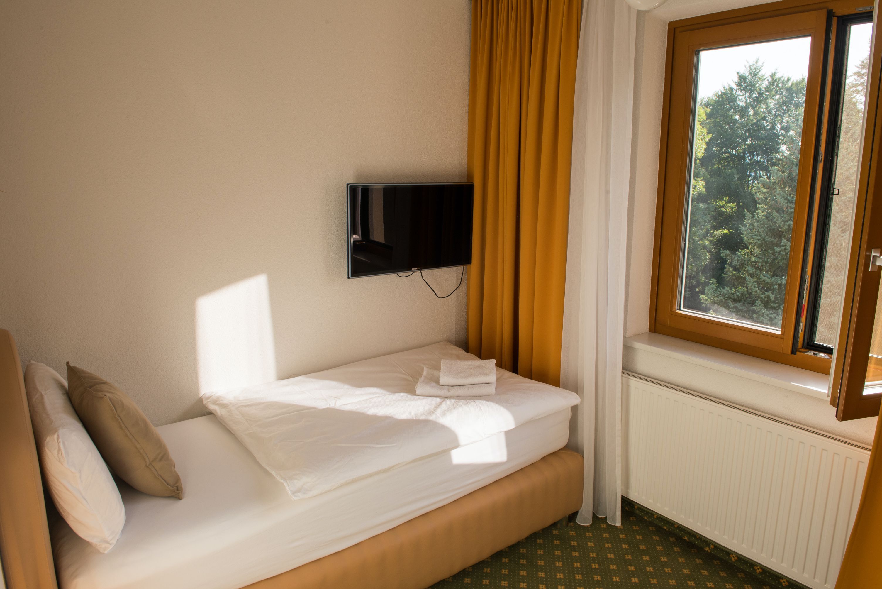 Jednolôžková izba s jednou posteľou, žltými závesmi, oknom s výhľadom na zeleň a nástennou TV.