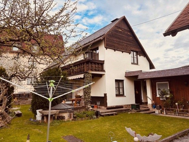 Dvojpodlažný dom s balkónom a záhradou, obklopený stromami a sušiakom v popredí.