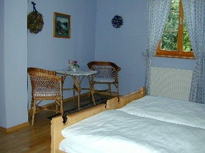 Útulná izba s dvoma posteľami, malým stolom a dvoma stoličkami. Viditeľné sú okná so závesmi a nástenné dekorácie.