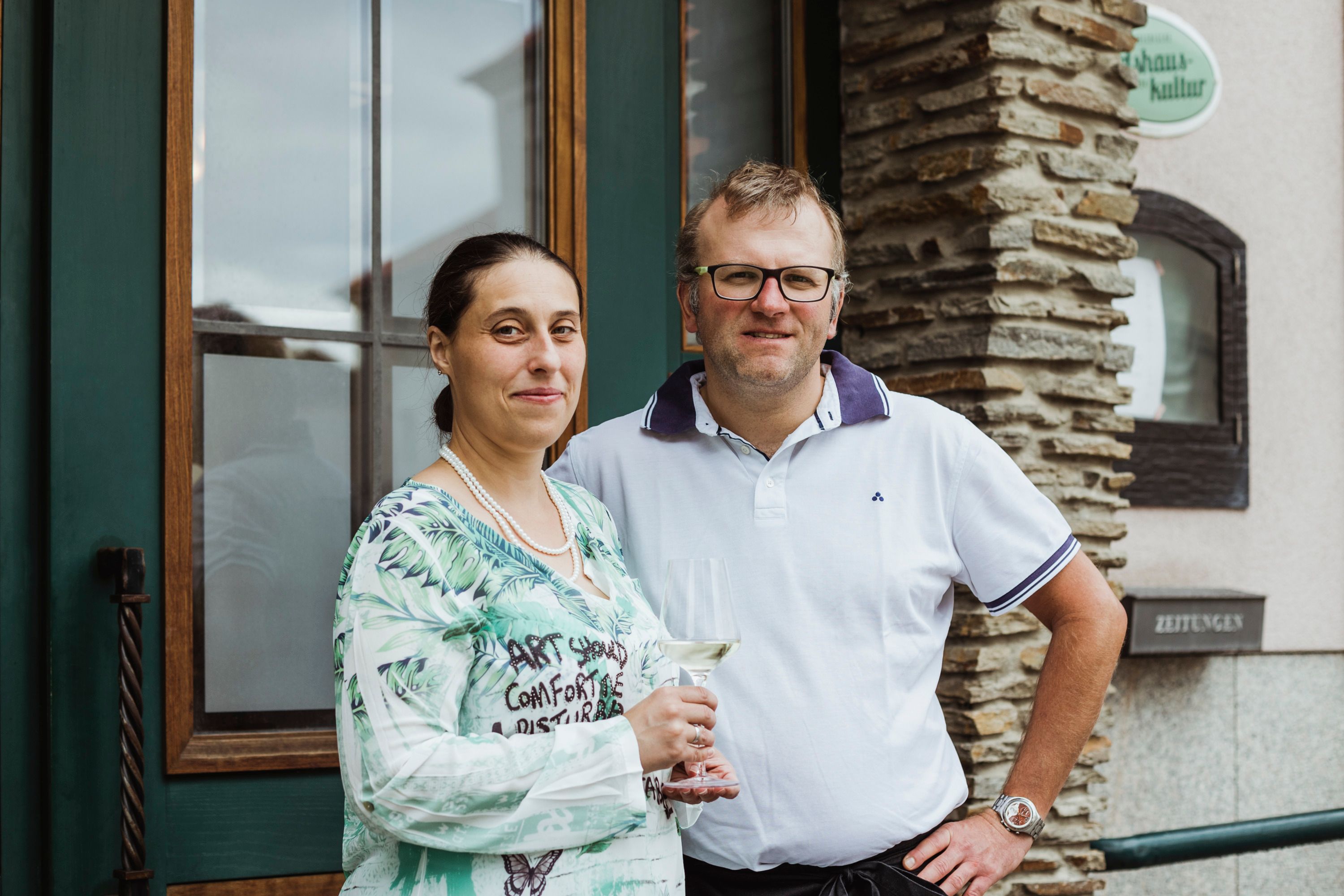 Muž a žena stoja s úsmevom pred budovou. Žena drží pohár s vínom.