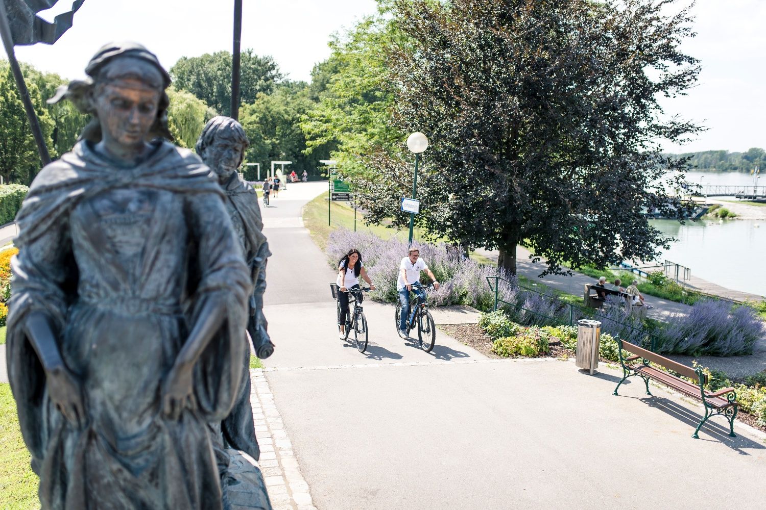 Dvaja ľudia jazdia na bicykli okolo sochy na Cyklotrasách obklopených stromami a riekou v pozadí.