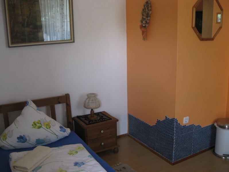 Malá izba s jednou posteľou, nočným stolíkom, lampou a oranžovou stenou s mozaikovým obkladom.
