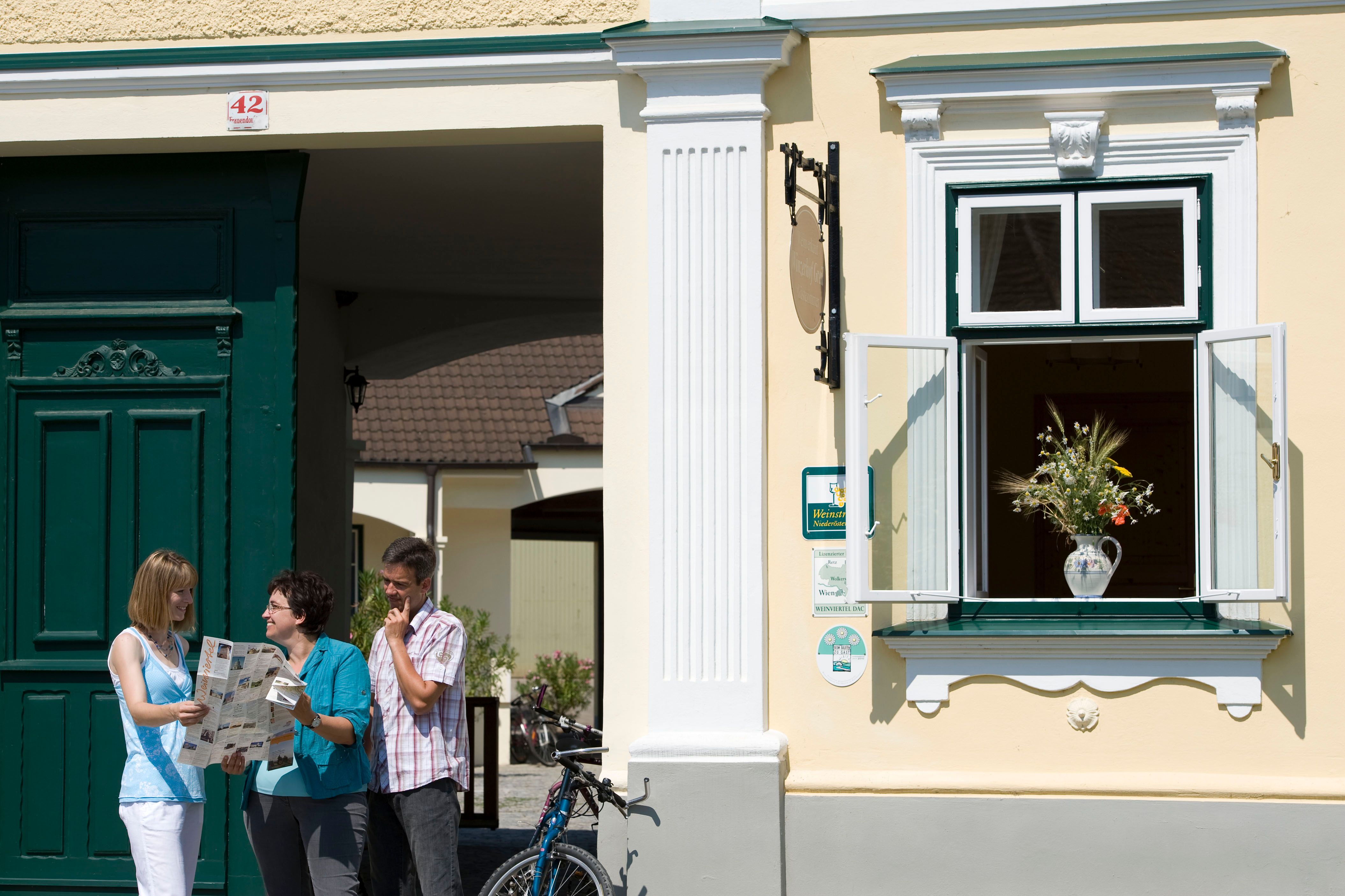 Pred žltou budovou so zelenými dverami stoja traja ľudia a čítajú si mapu. Okno s kyticou kvetov je otvorené.