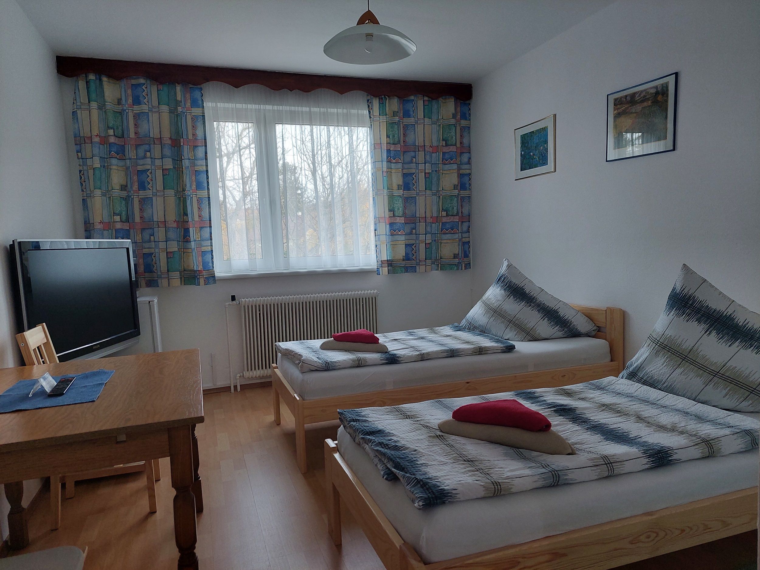 Jednoducho zariadená izba s dvoma samostatnými posteľami, stolíkom, TV a oknom s farebnými závesmi.