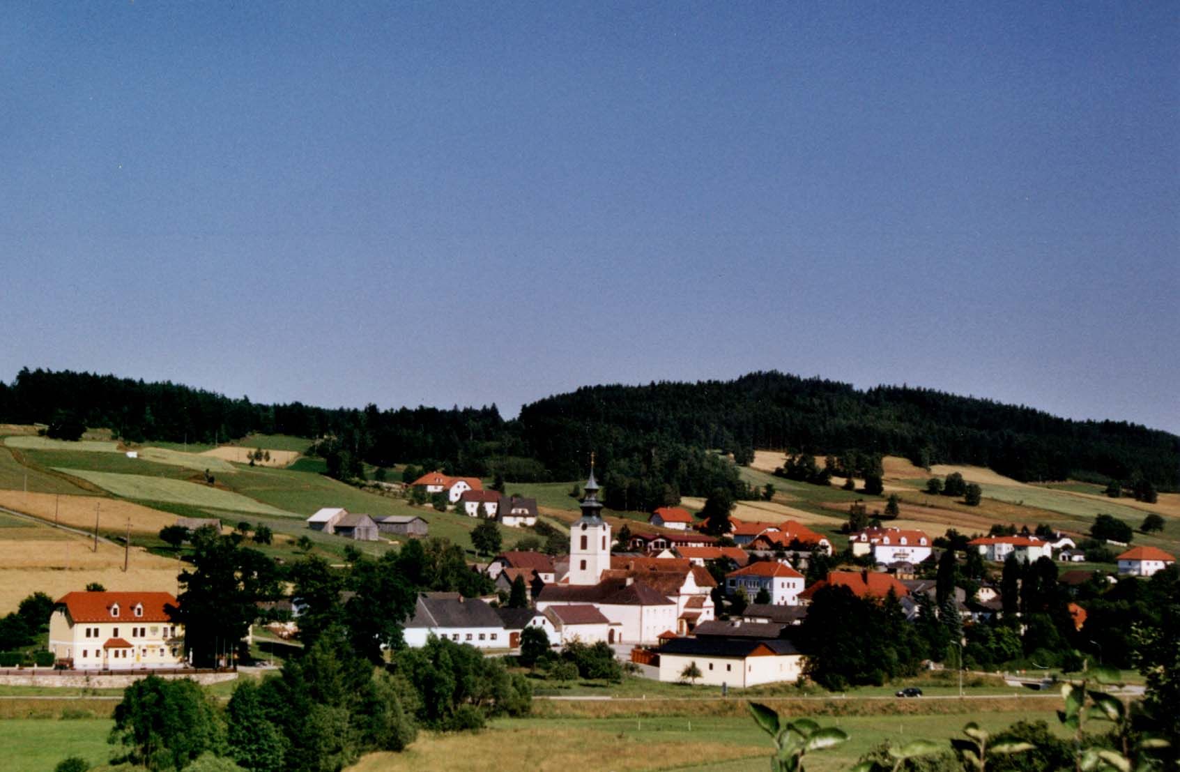 Panoráma dediny s kostolom a červenými strechami, obklopenej zelenými poliami a lesmi.
