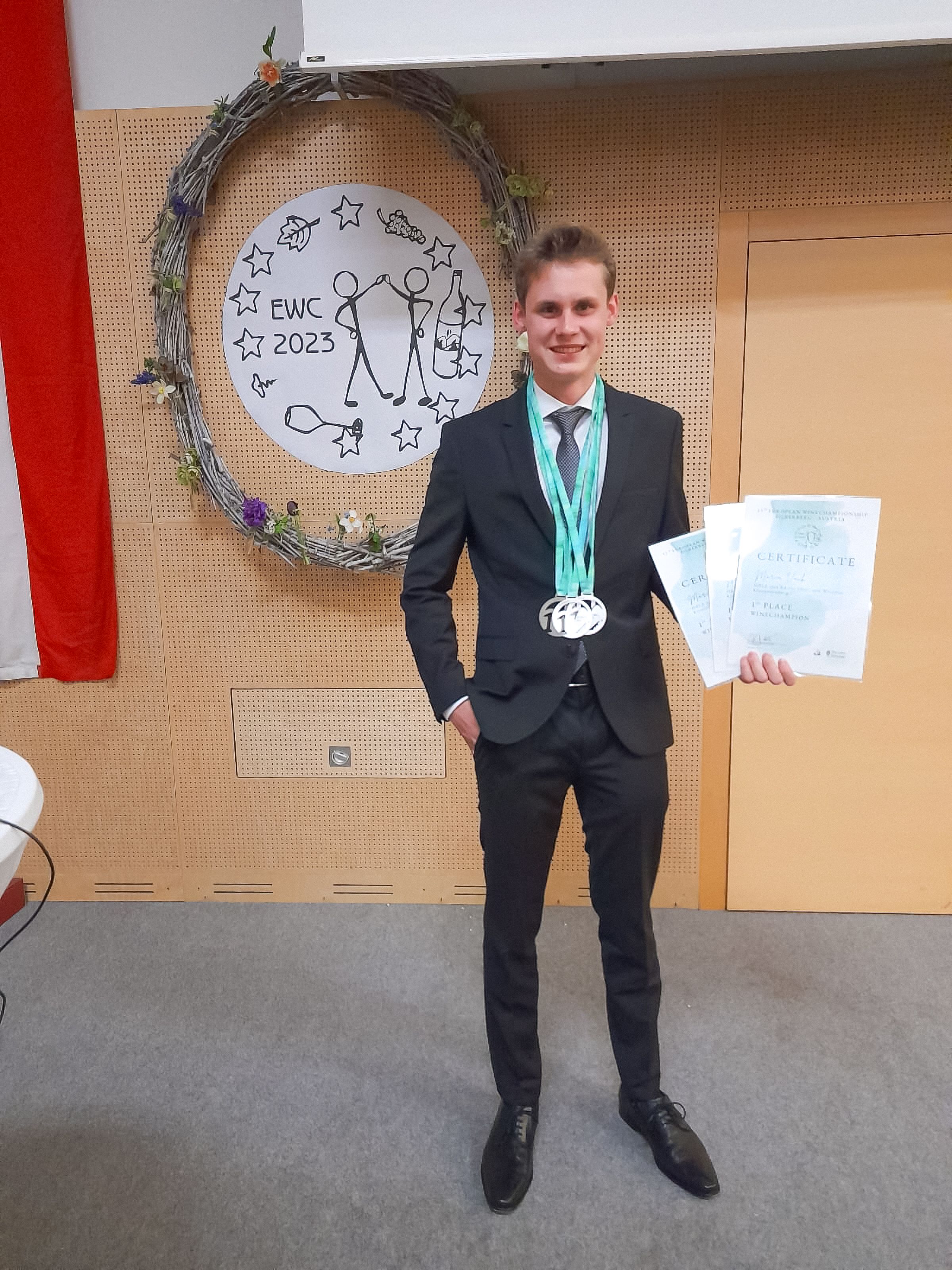 Mladý muž v obleku stojí pred ozdobným vencom s certifikátom a medailami v ruke.