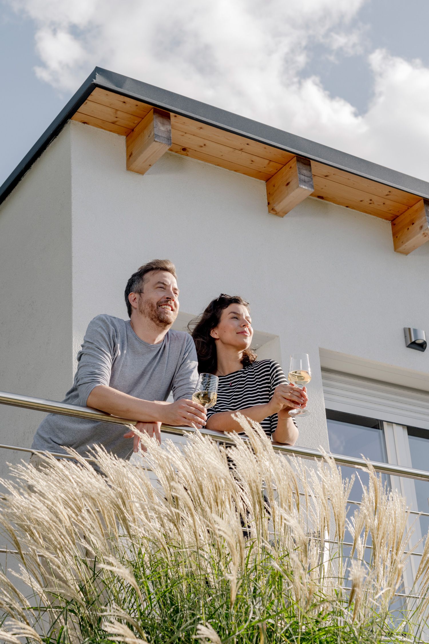Pár stojí na balkóne s pohármi vína a v pozadí je moderná budova.