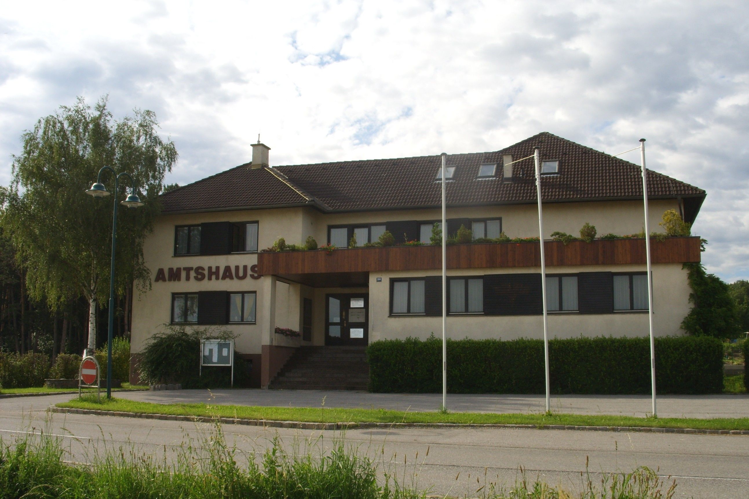 Administratívna budova Natschbach-Loipersbach s hnedou strechou a balkónom, obklopená stromami a stožiarmi.