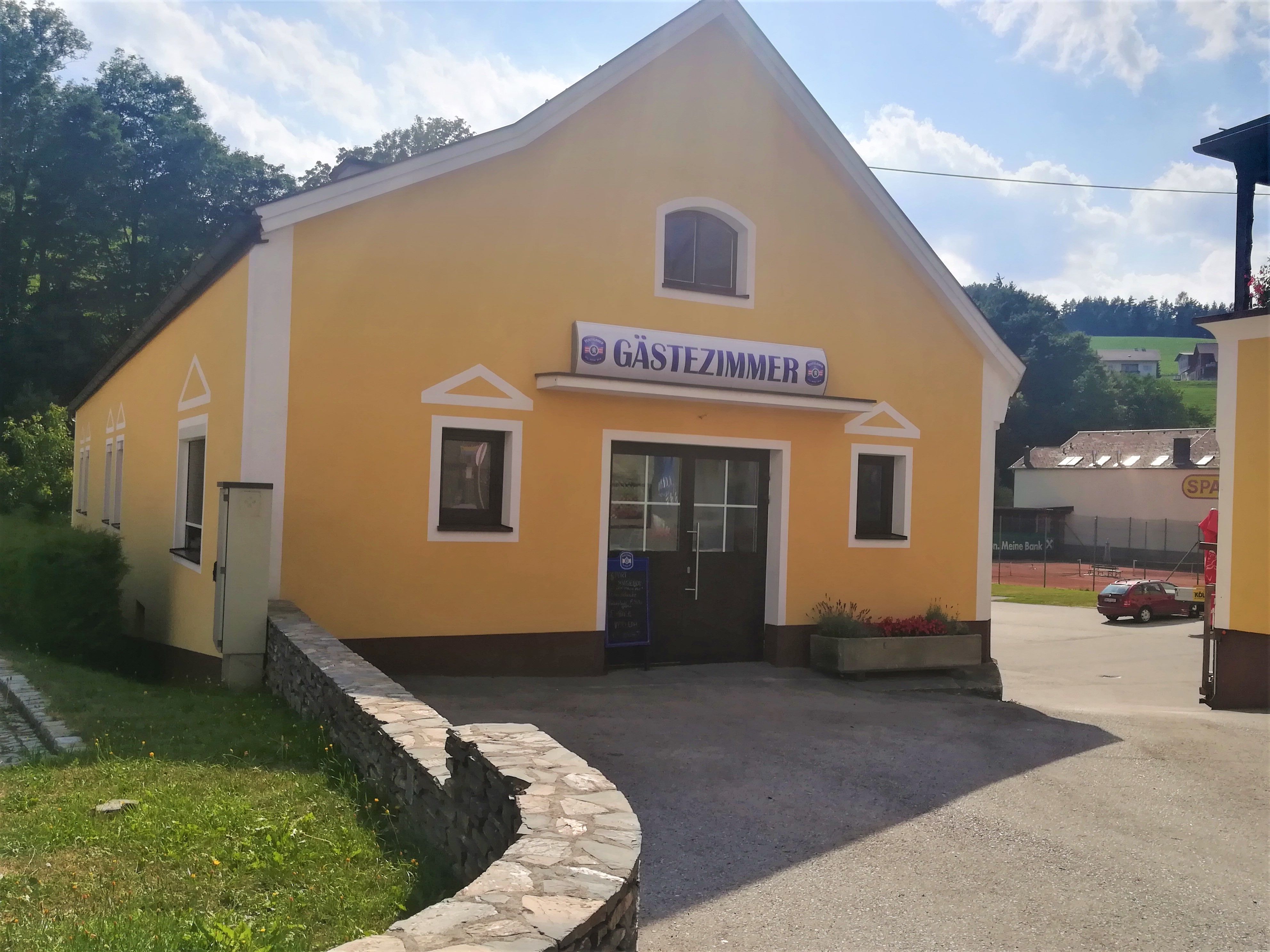 Žltá budova s nápisom "izba pre hostí", obklopená stromami a parkoviskom.